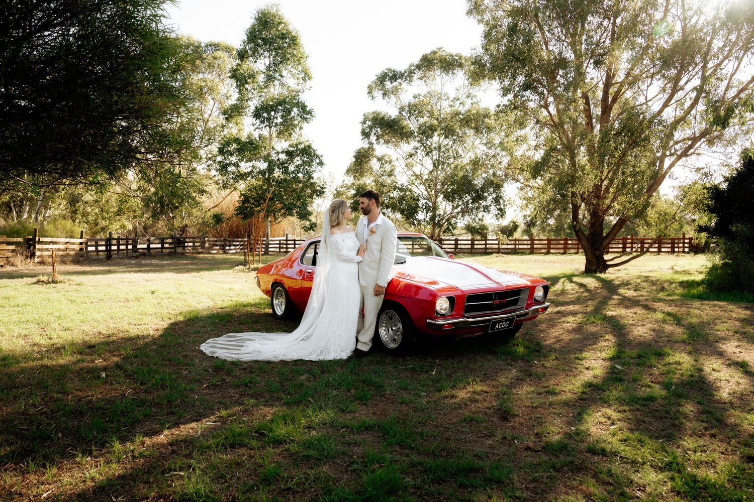 Bride and groom pose beside a classic red Mustang on a sunny day at a rustic outdoor wedding venue.