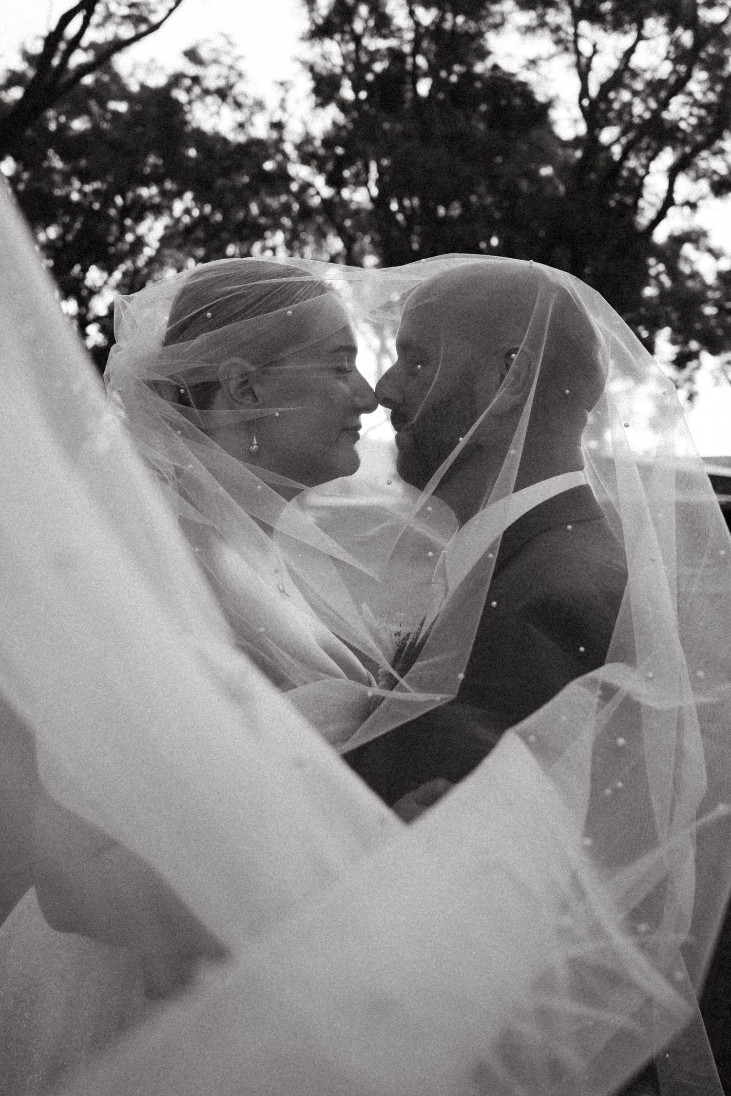 Black and white wedding photo of bride and groom sharing an intimate moment under a flowing veil outdoors.
