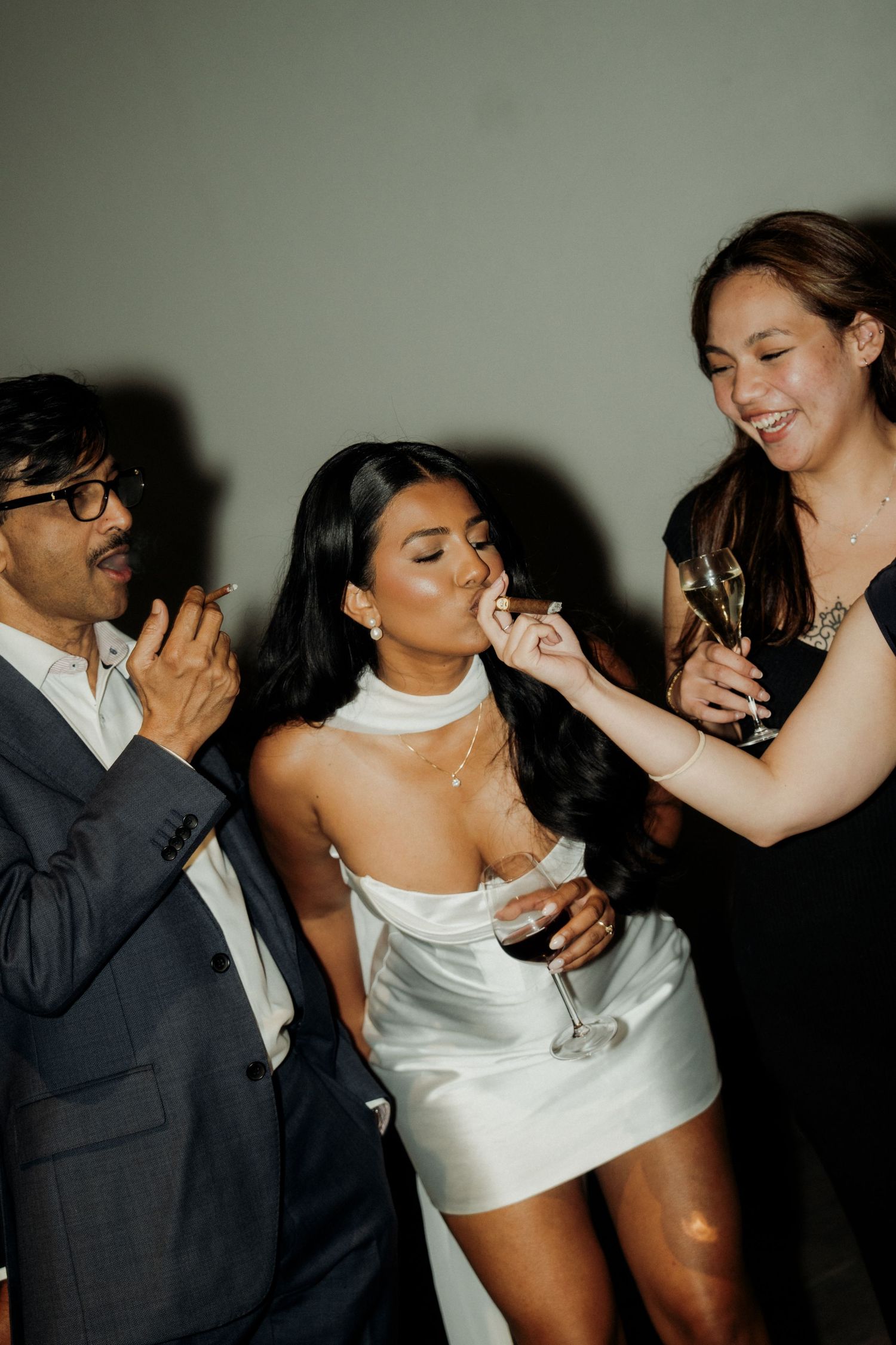 Three guests celebrating at a party, woman in white dress sipping a drink while friends in formal attire smile nearby.