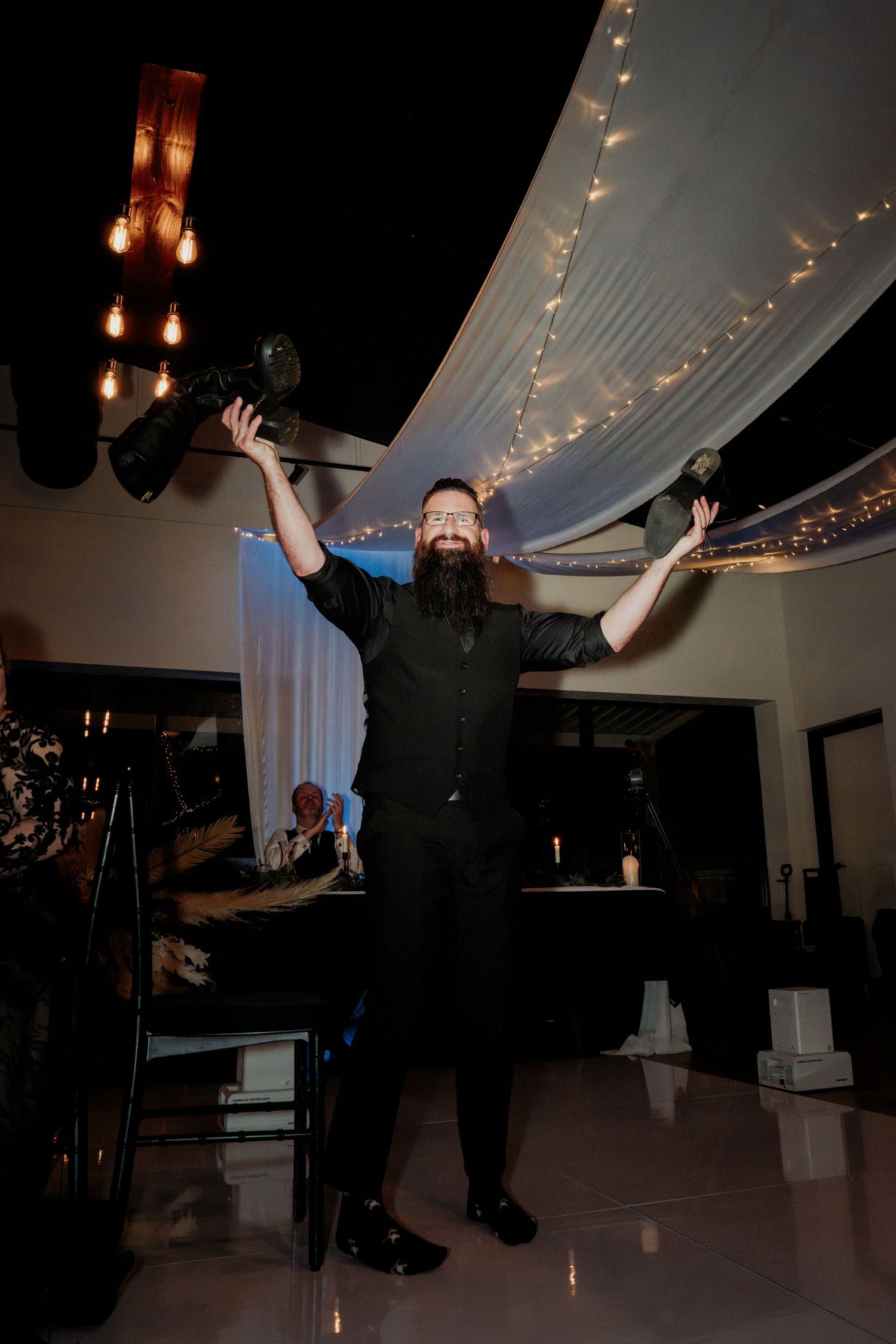 Bearded man in black outfit triumphantly raises two bottles at an elegant event venue with fairy lights.