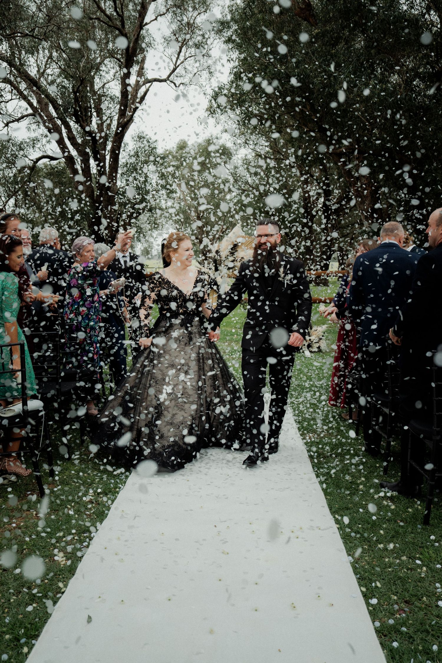 Bride in black wedding dress and groom walk down aisle through confetti shower outdoors.