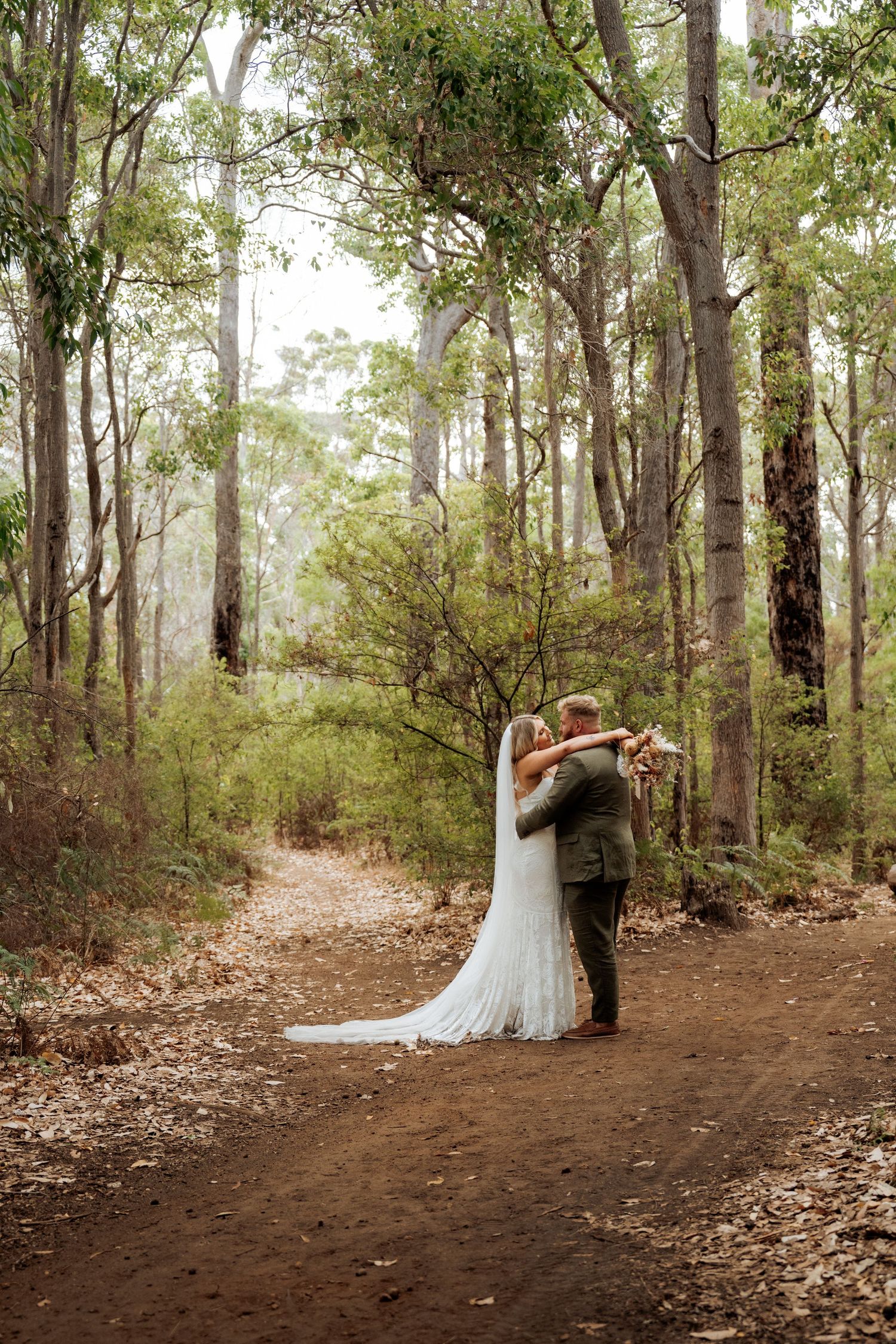 Bride and groom embracing on a forest path surrounded by tall eucalyptus trees in a romantic woodland wedding setting.