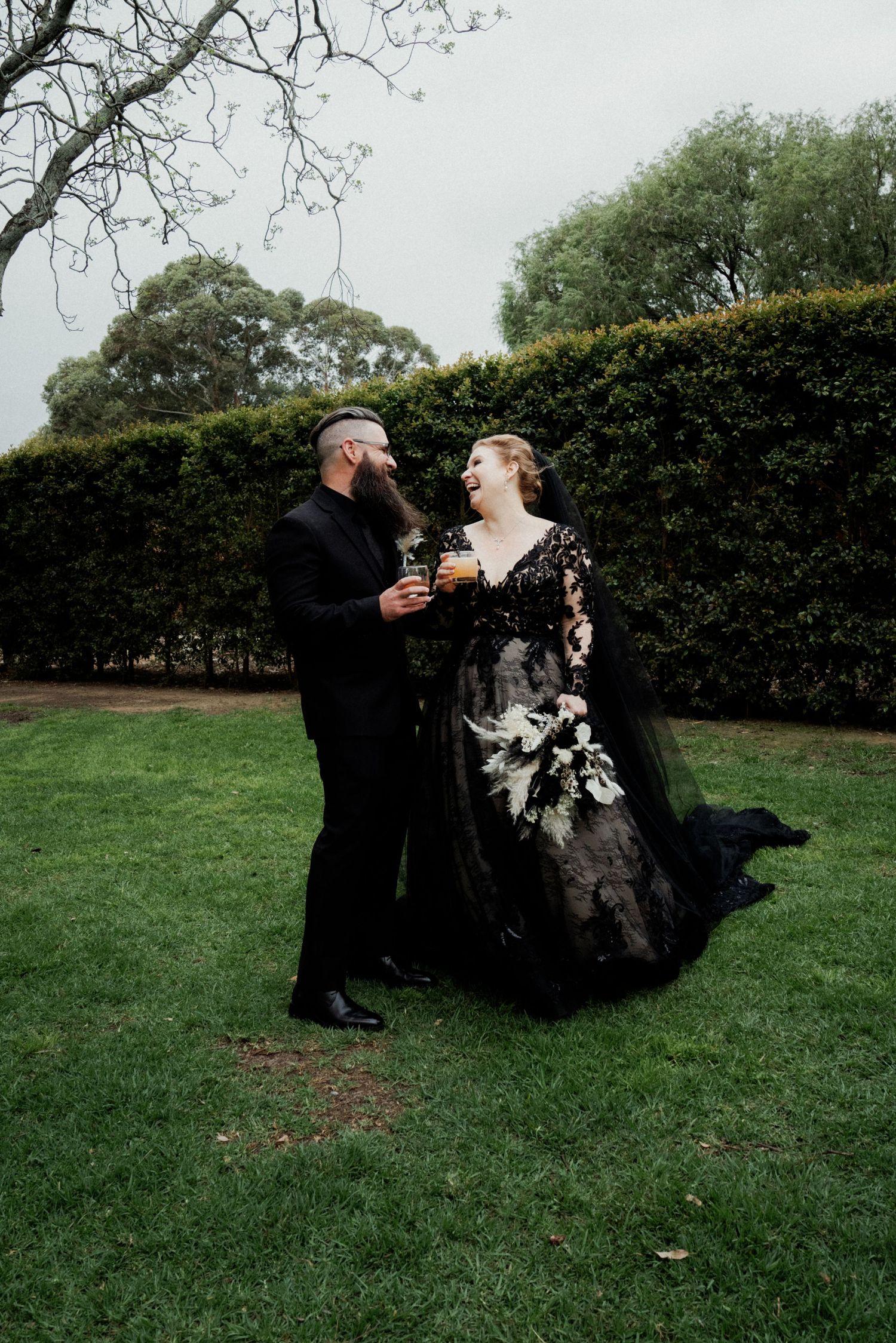 Bride in black lace gown and groom in black suit toast drinks outdoors against green hedges.