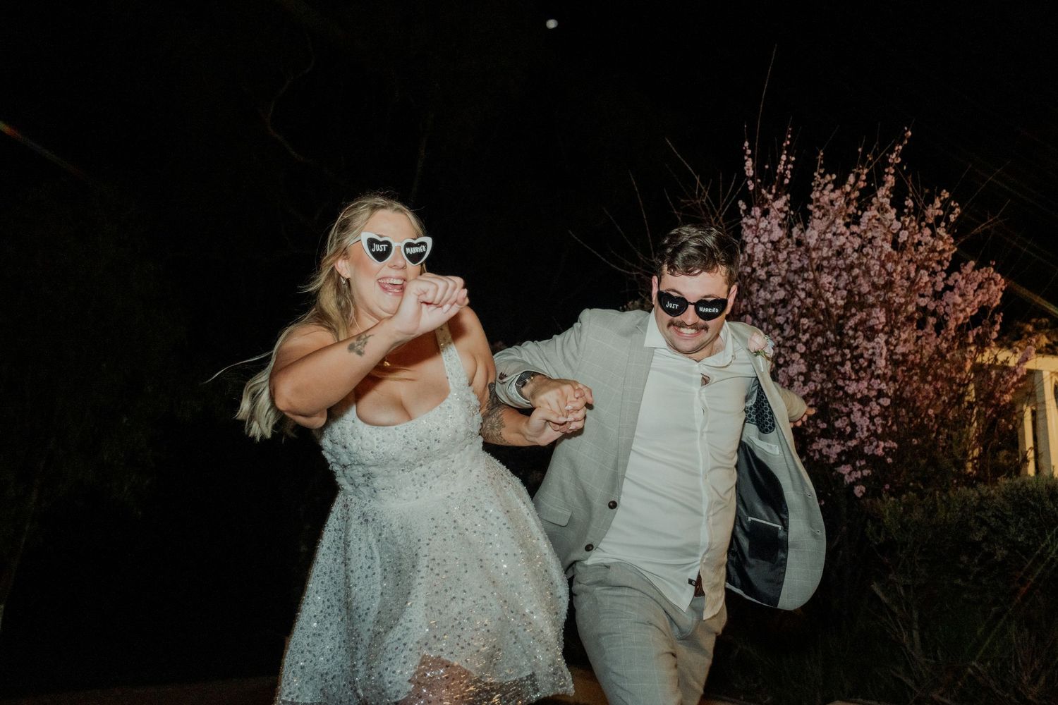 Bride and groom wearing fun sunglasses laughing together outdoors at night during wedding celebration.