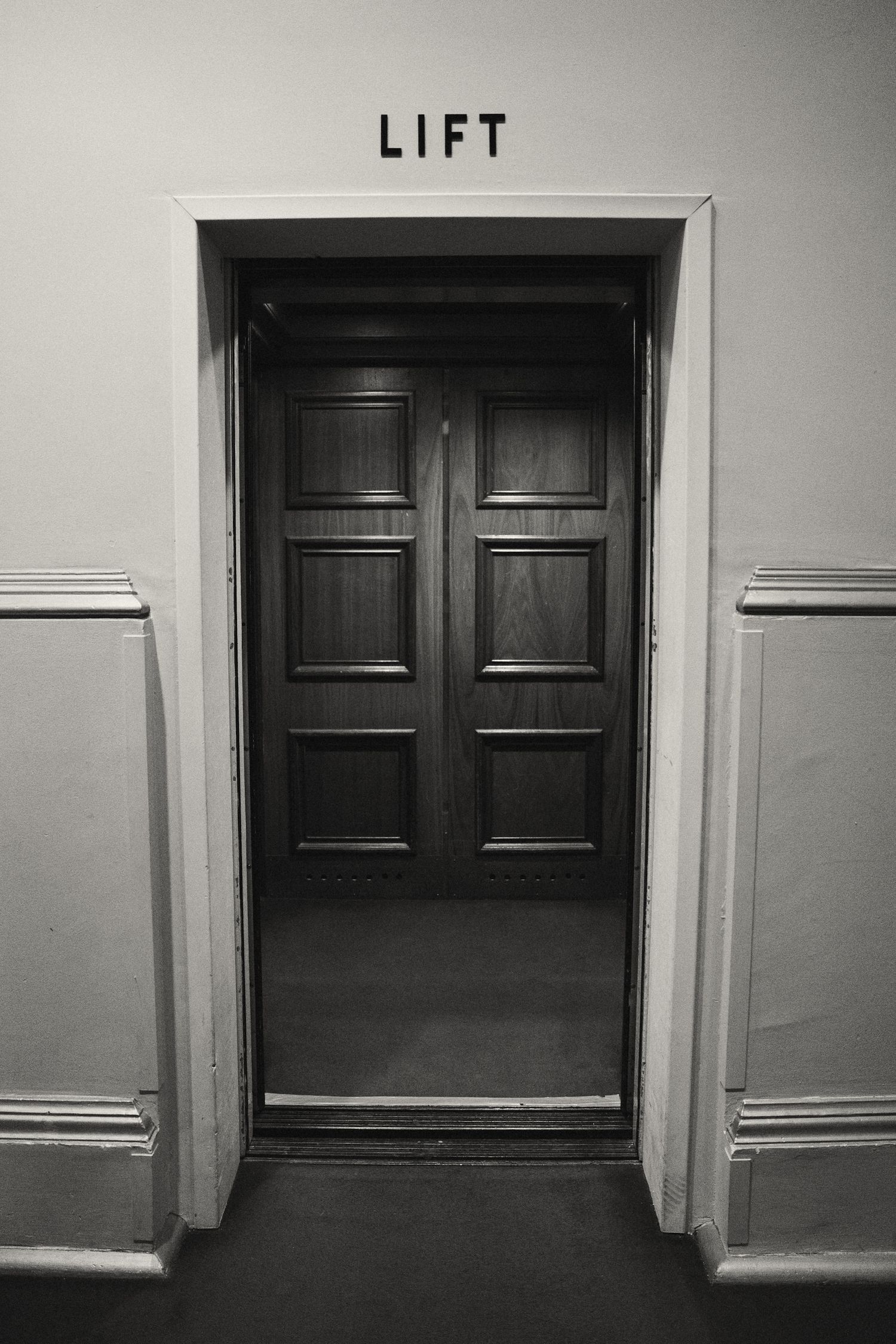 A black and white photograph of an open elevator entrance with a simple 'LIFT' sign above dark wooden doors.