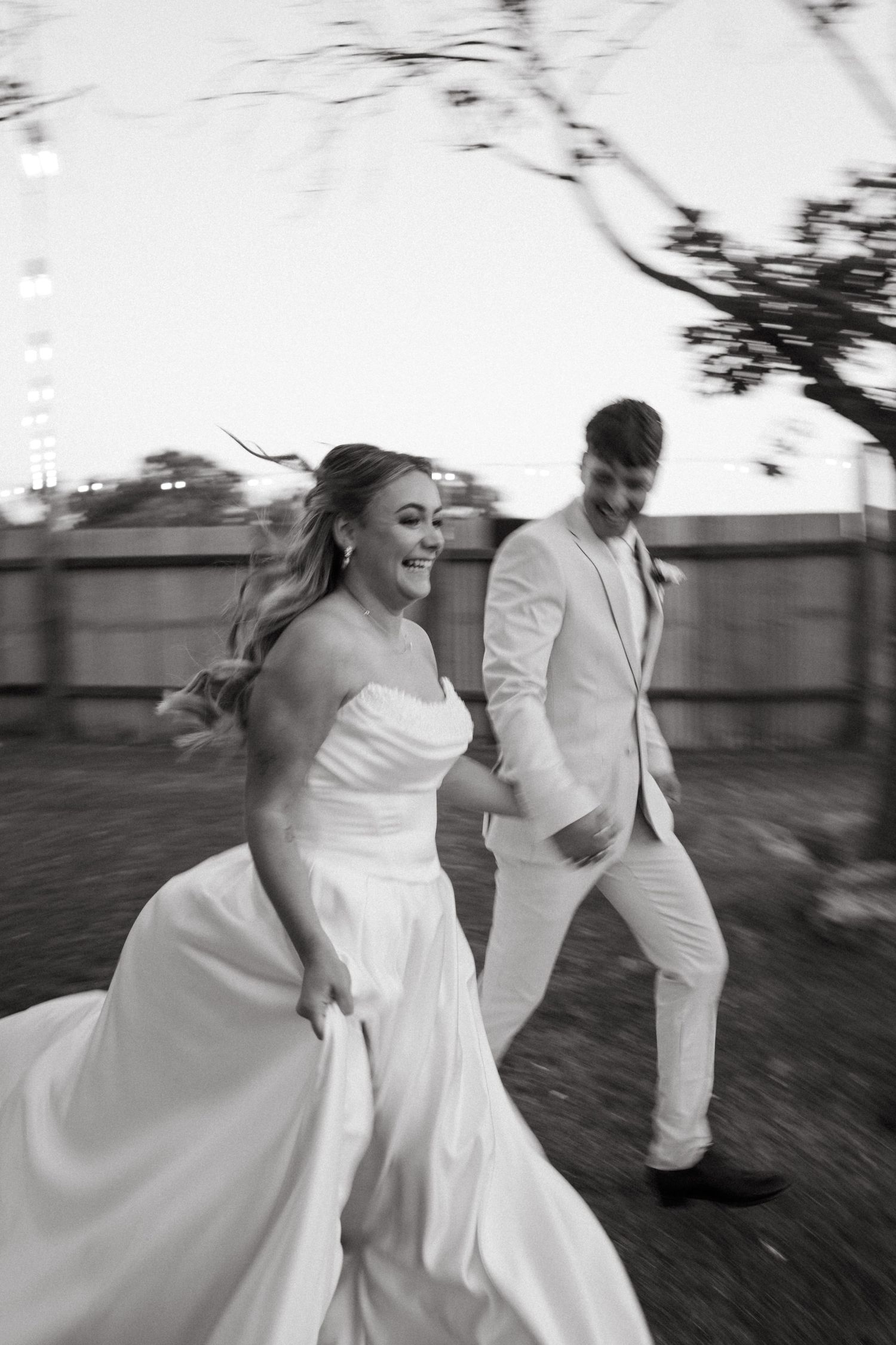 Black and white photo of a joyful bride and groom running together outdoors on their wedding day.