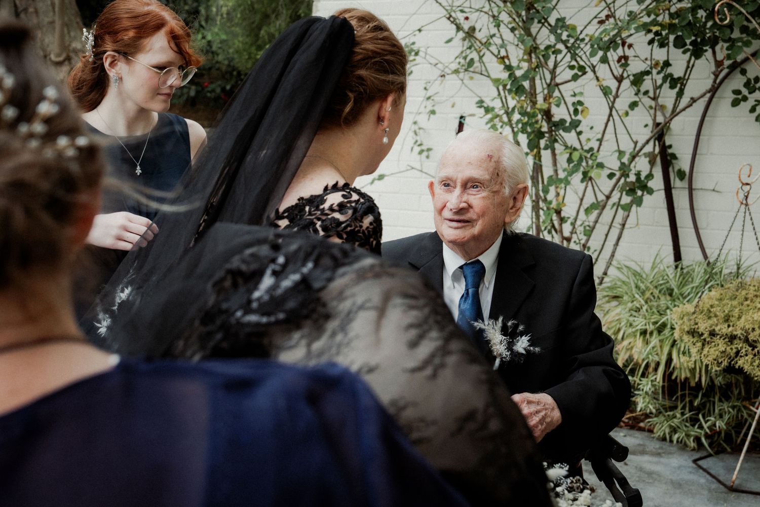 Elderly man in suit with boutonniere converses with women in black lace dresses at a wedding reception.
