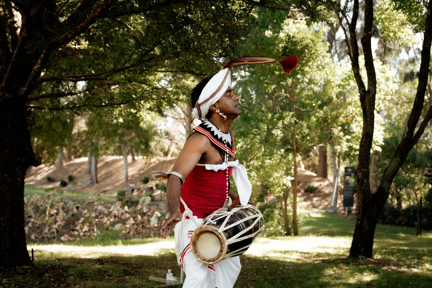 Traditional dancer in red costume plays a drum outdoors among green trees in a cultural performance.