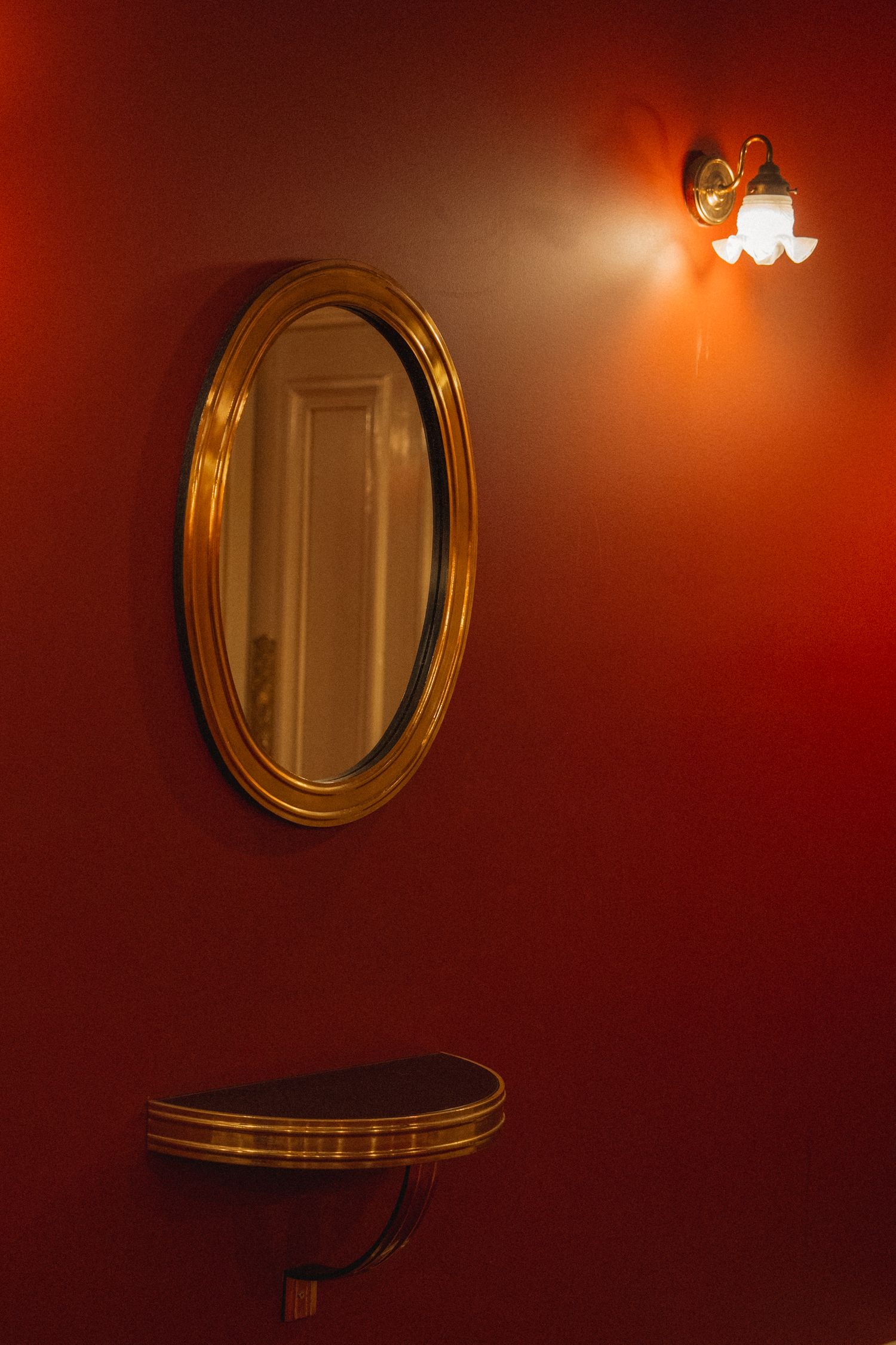 An oval gold-framed mirror hangs above a matching half-moon table on a deep red wall with a vintage wall sconce.