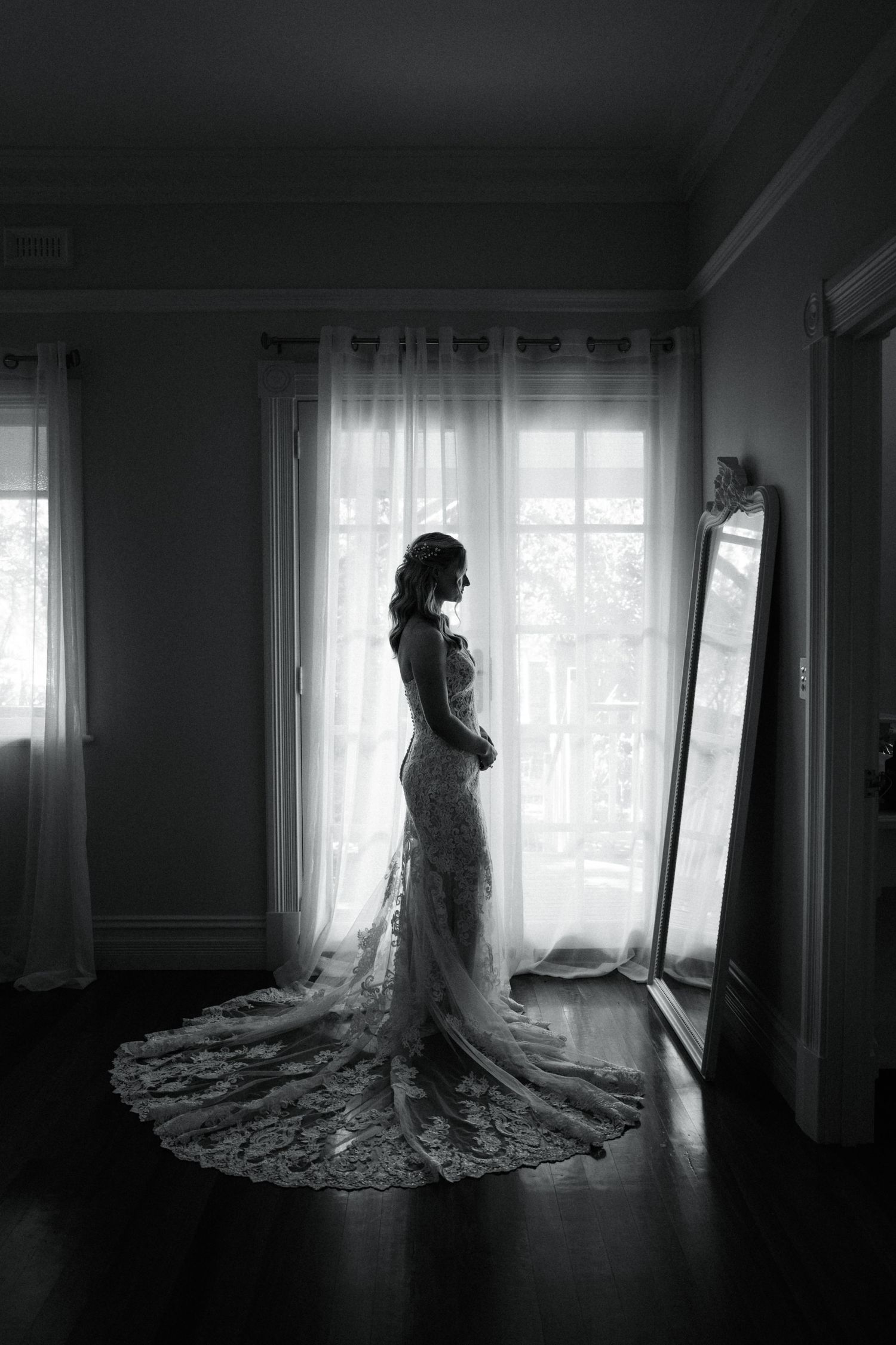 Black and white photo of a bride in a lace wedding gown standing by a sunlit window with a mirror nearby.