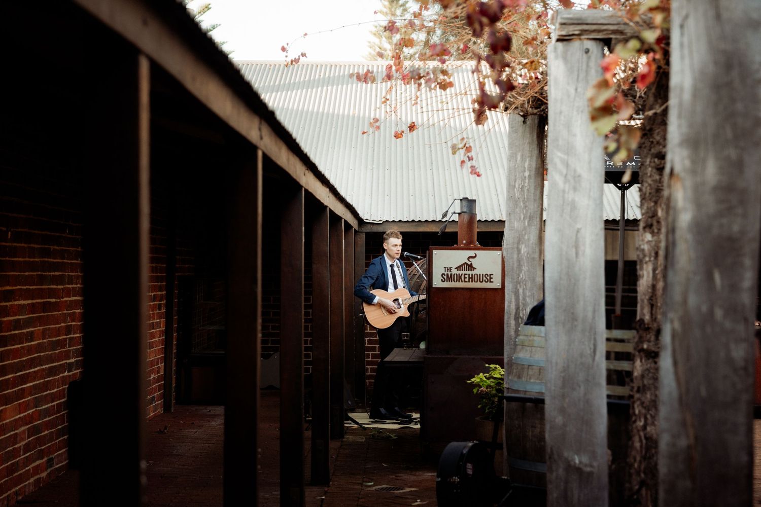 Man playing acoustic guitar in a rustic brick courtyard with autumn foliage and corrugated iron roofing.