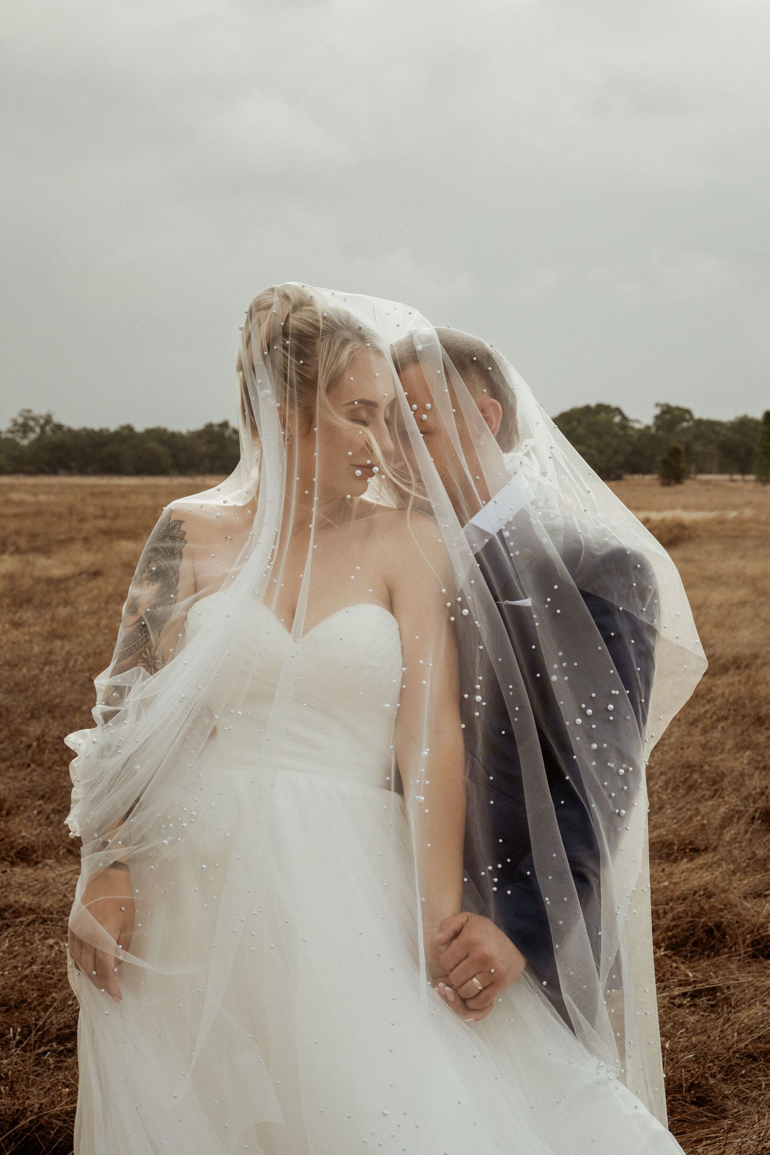 Bride and groom share a romantic moment under a pearl-adorned veil in an open field on their wedding day.
