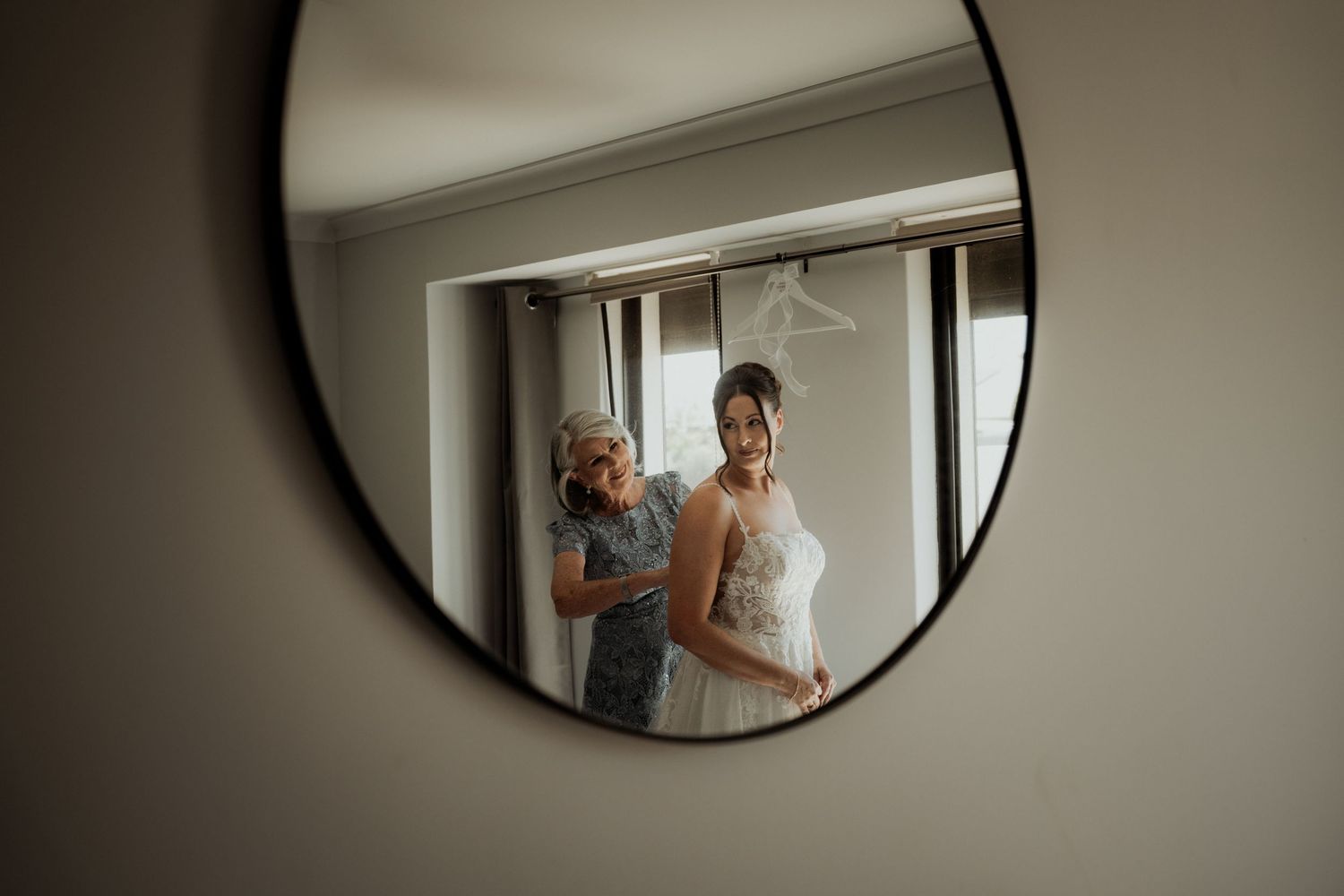 Bride getting dressed with help reflected in a round mirror, wearing a white wedding gown before the ceremony.
