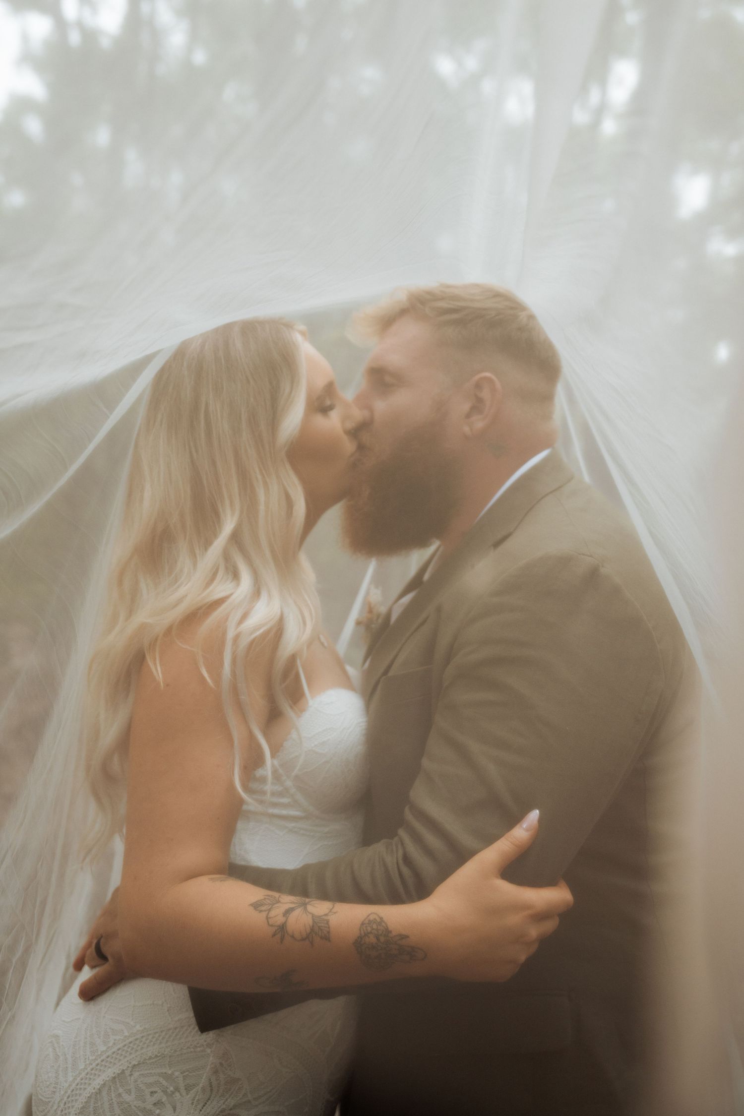 Bride and groom sharing a romantic kiss under a flowing wedding veil, surrounded by soft natural light.