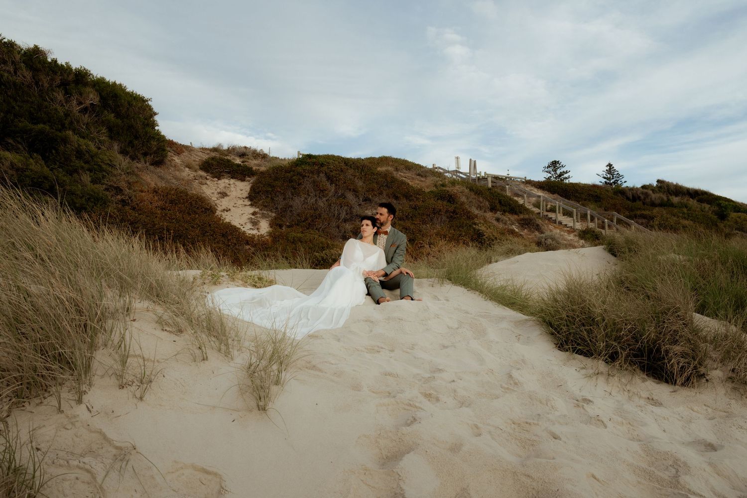 Bride and groom sitting together on sandy beach dunes with flowing white wedding dress, surrounded by coastal grass.