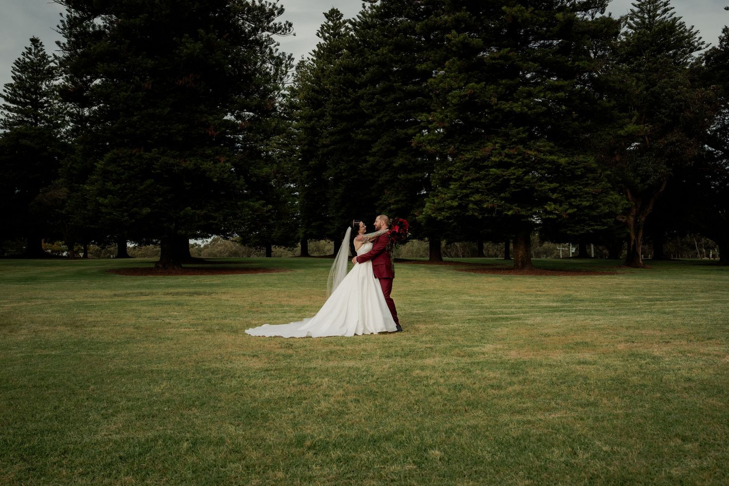 Bride and groom share a romantic moment on a lush green lawn surrounded by tall trees on their wedding day.