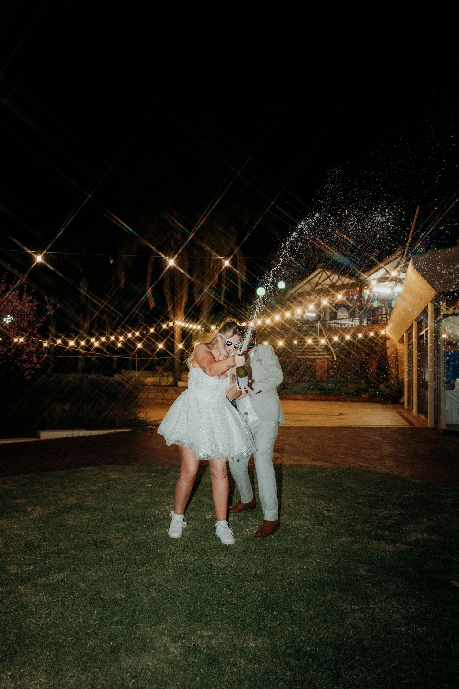 Bride and groom celebrating outdoors at night, popping champagne under string lights at a romantic wedding venue.