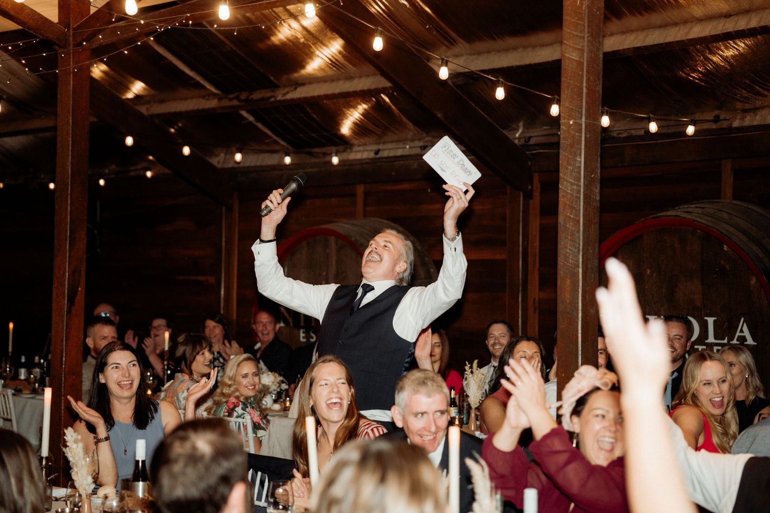 Joyful wedding MC raises arms in celebration at rustic barn reception as excited guests cheer and applaud around decorated tables.