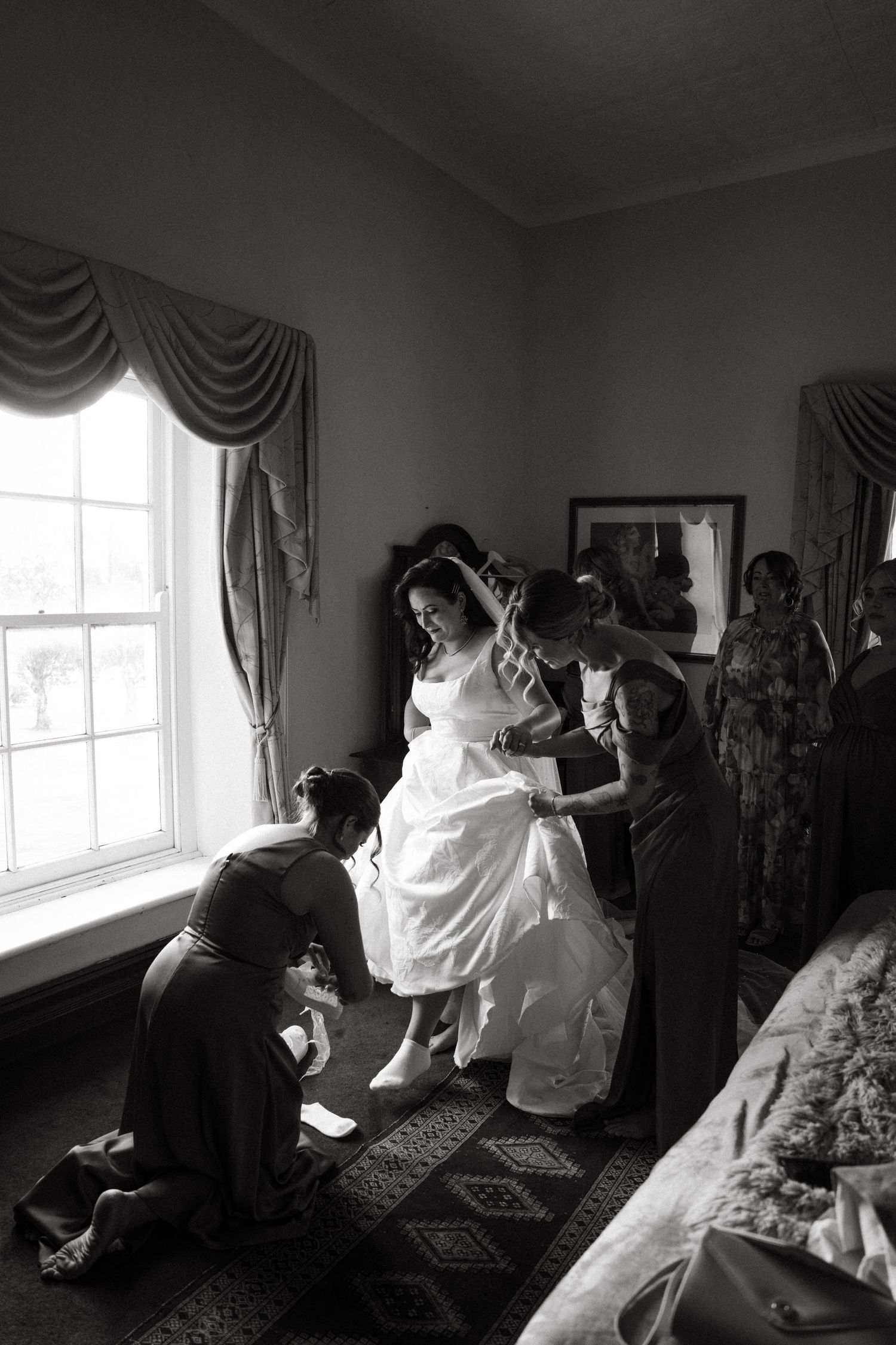 Black and white photo of bride being helped into wedding dress by bridesmaids near a window in a elegant room.