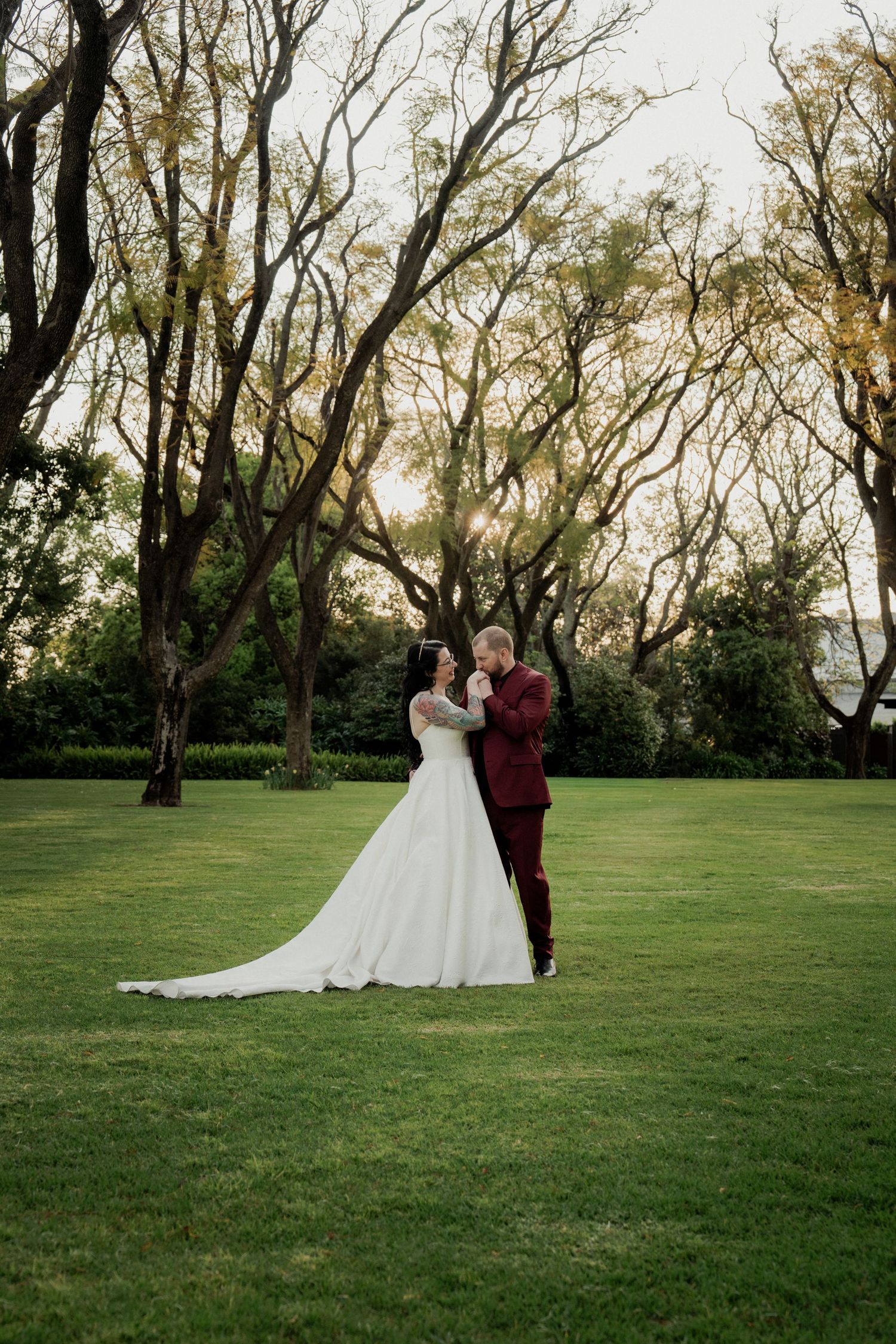 Bride and groom embrace on lush green lawn surrounded by tall trees at golden hour during outdoor wedding ceremony.