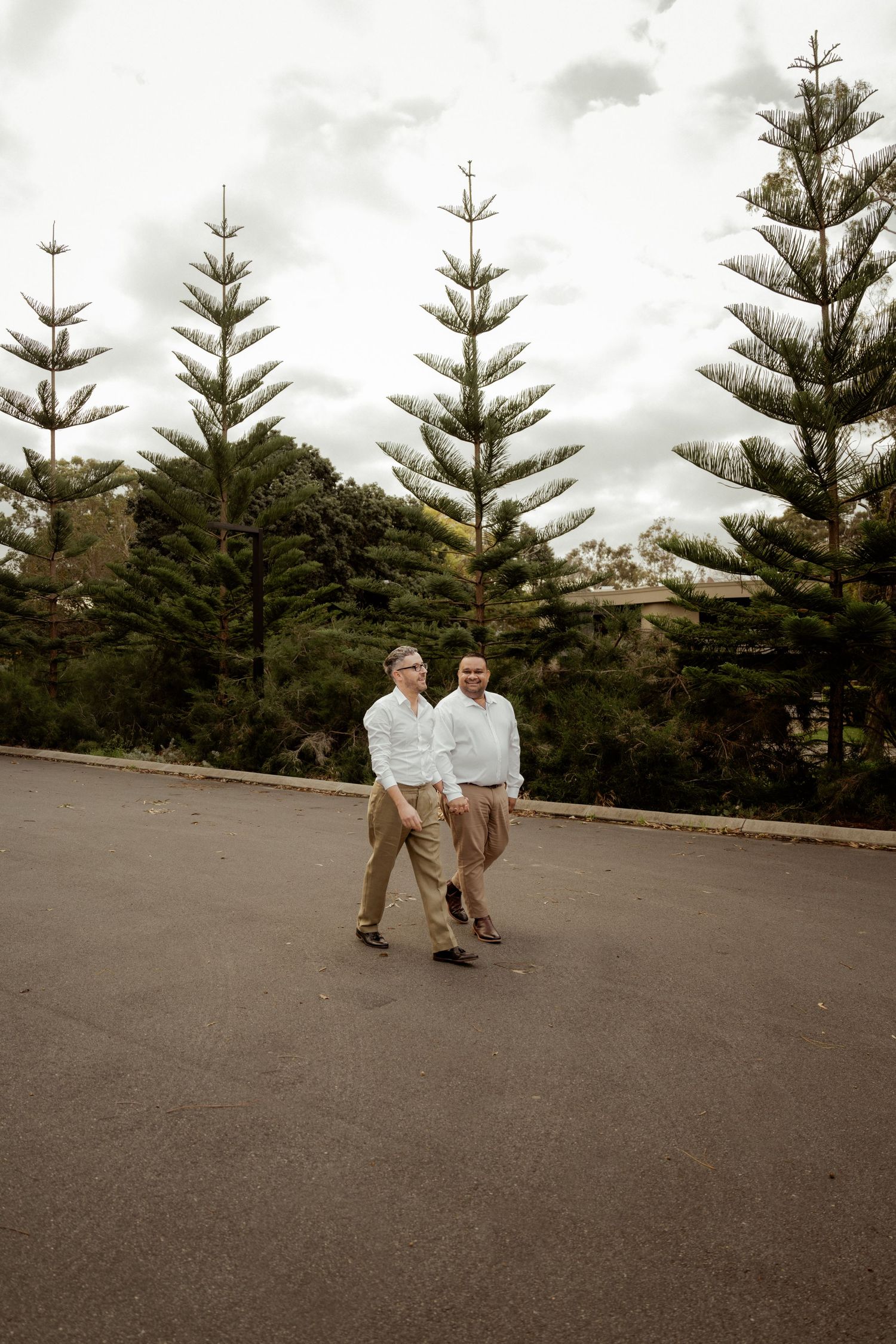 Two men in white shirts and khaki pants walk together on a paved path surrounded by tall pine trees.