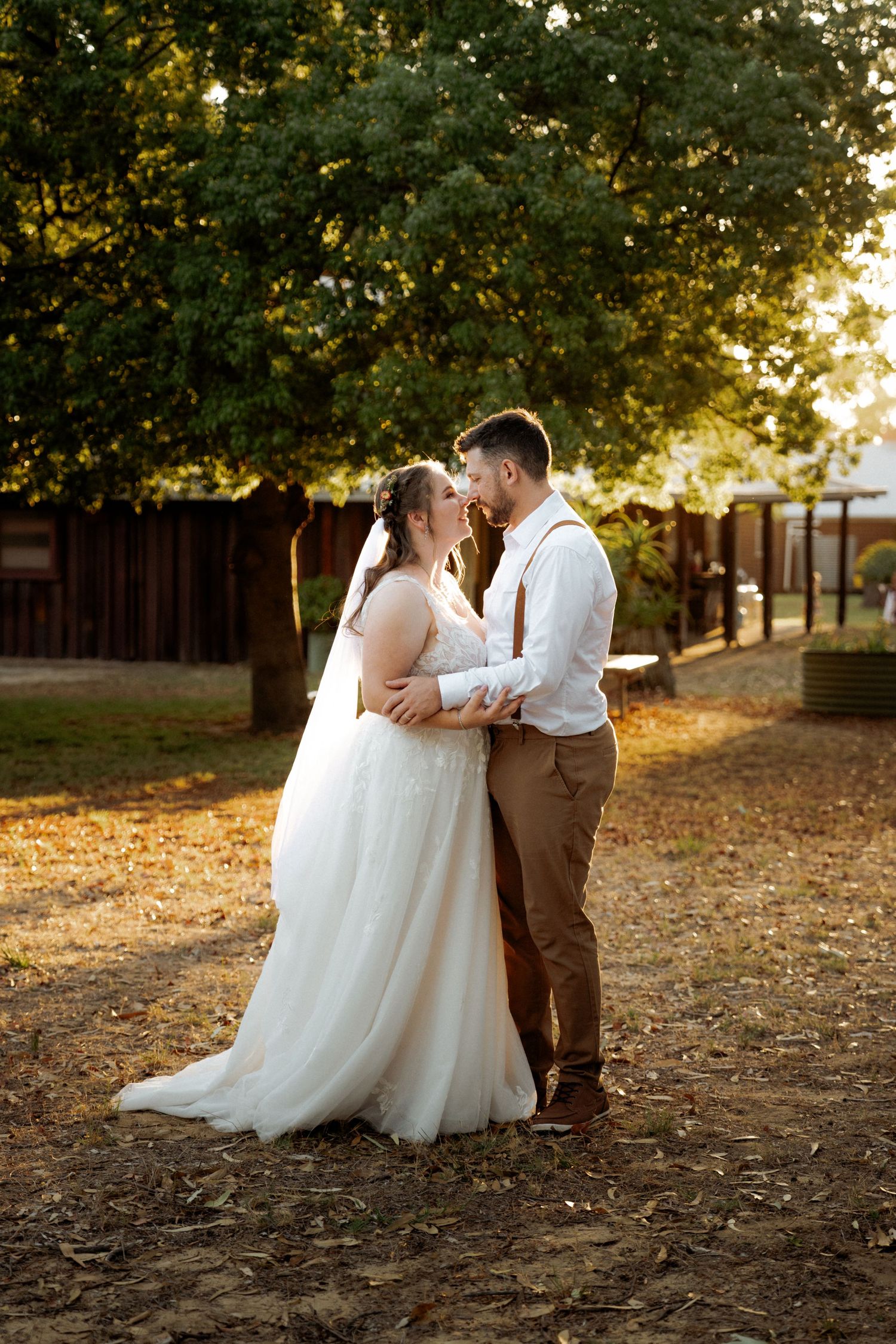 Bride and groom share a romantic moment outdoors at golden hour, surrounded by lush green trees at their wedding venue.
