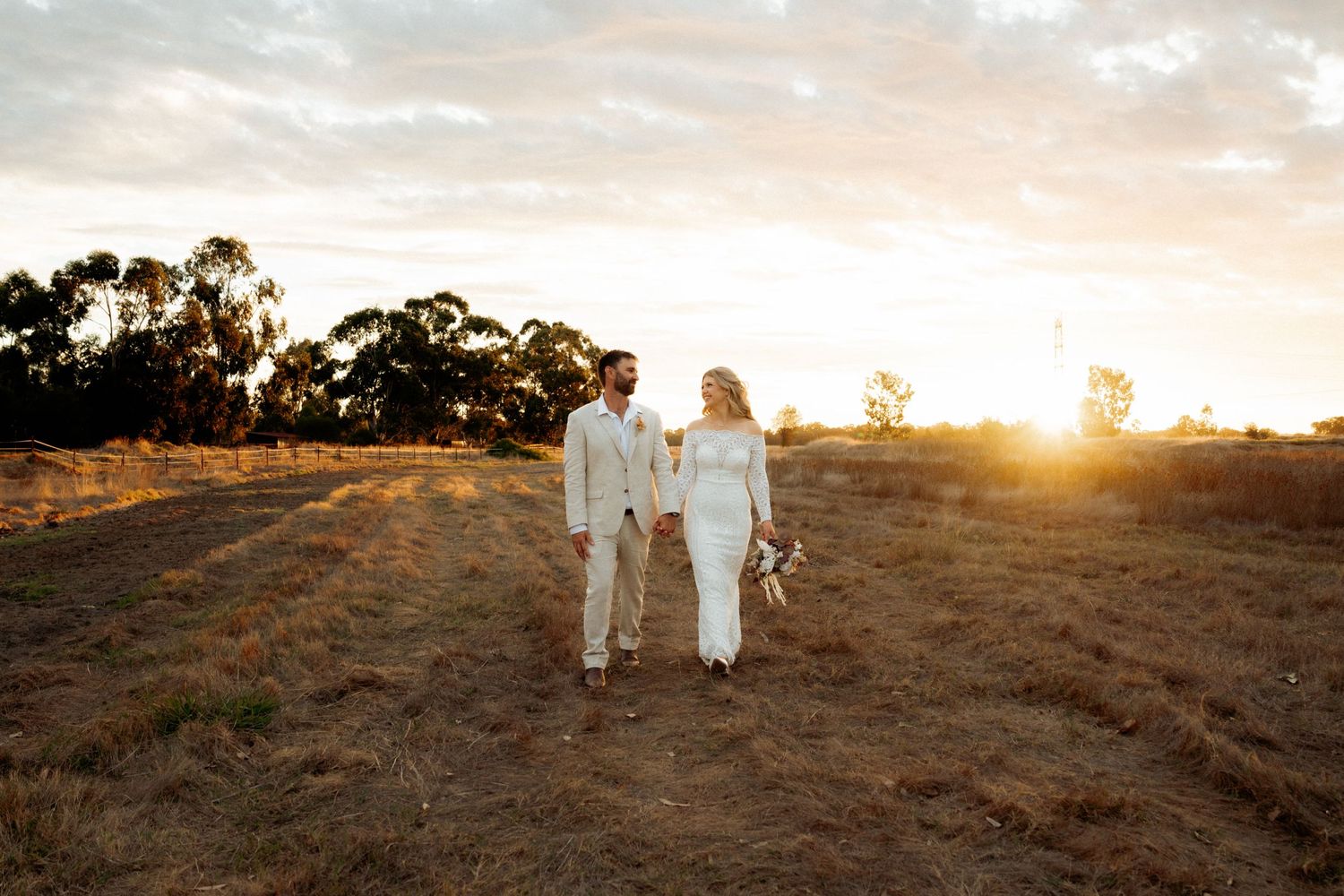 Bride and groom hold hands walking through golden field at sunset on their wedding day.