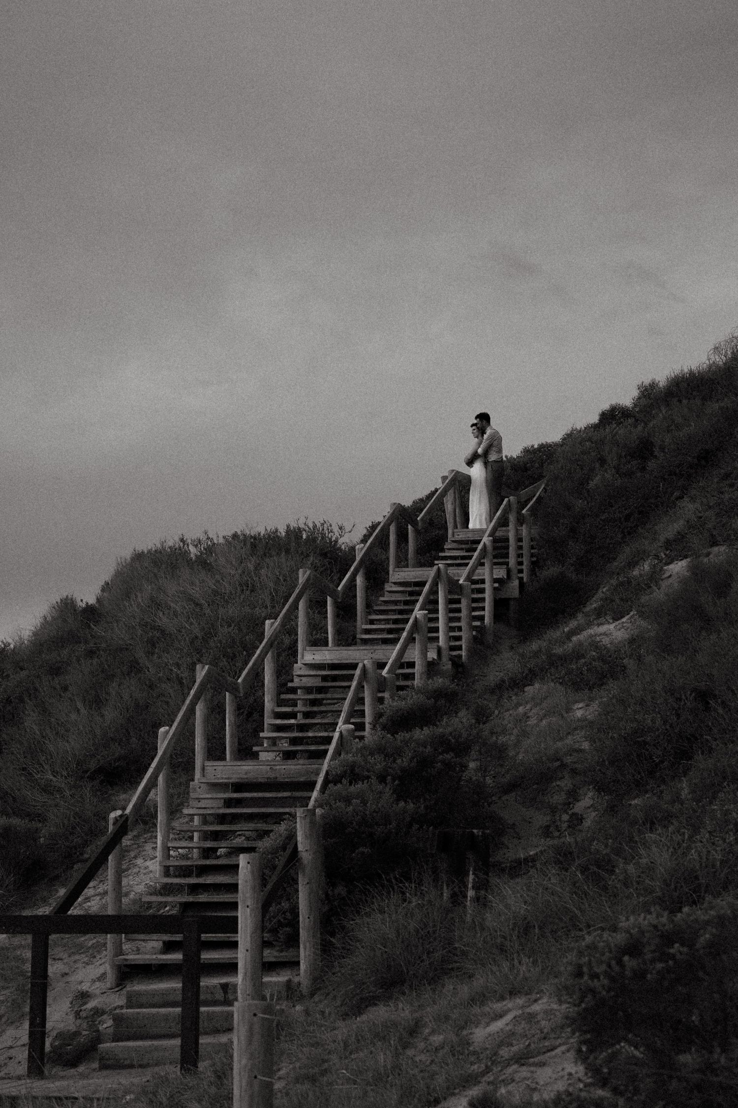 Black and white photo of a couple standing atop wooden beach stairs on a grassy hillside under cloudy skies.
