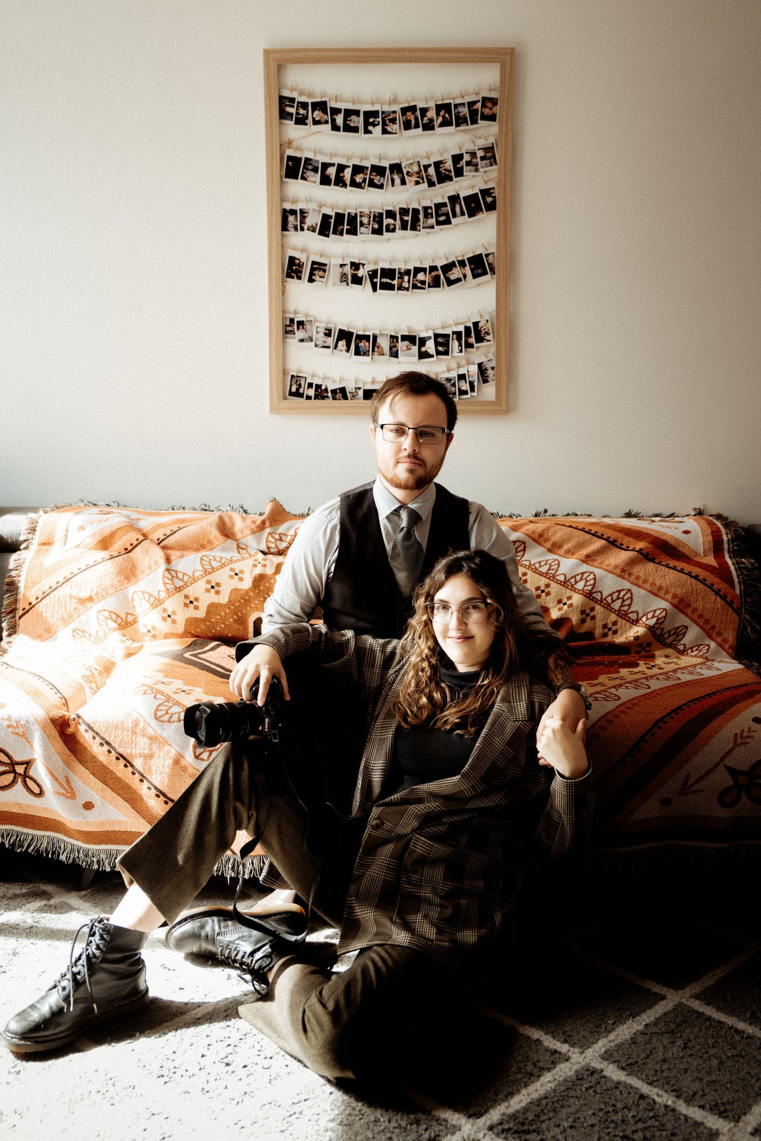 Couple in vintage attire sitting on orange patterned couch under hanging photographs.