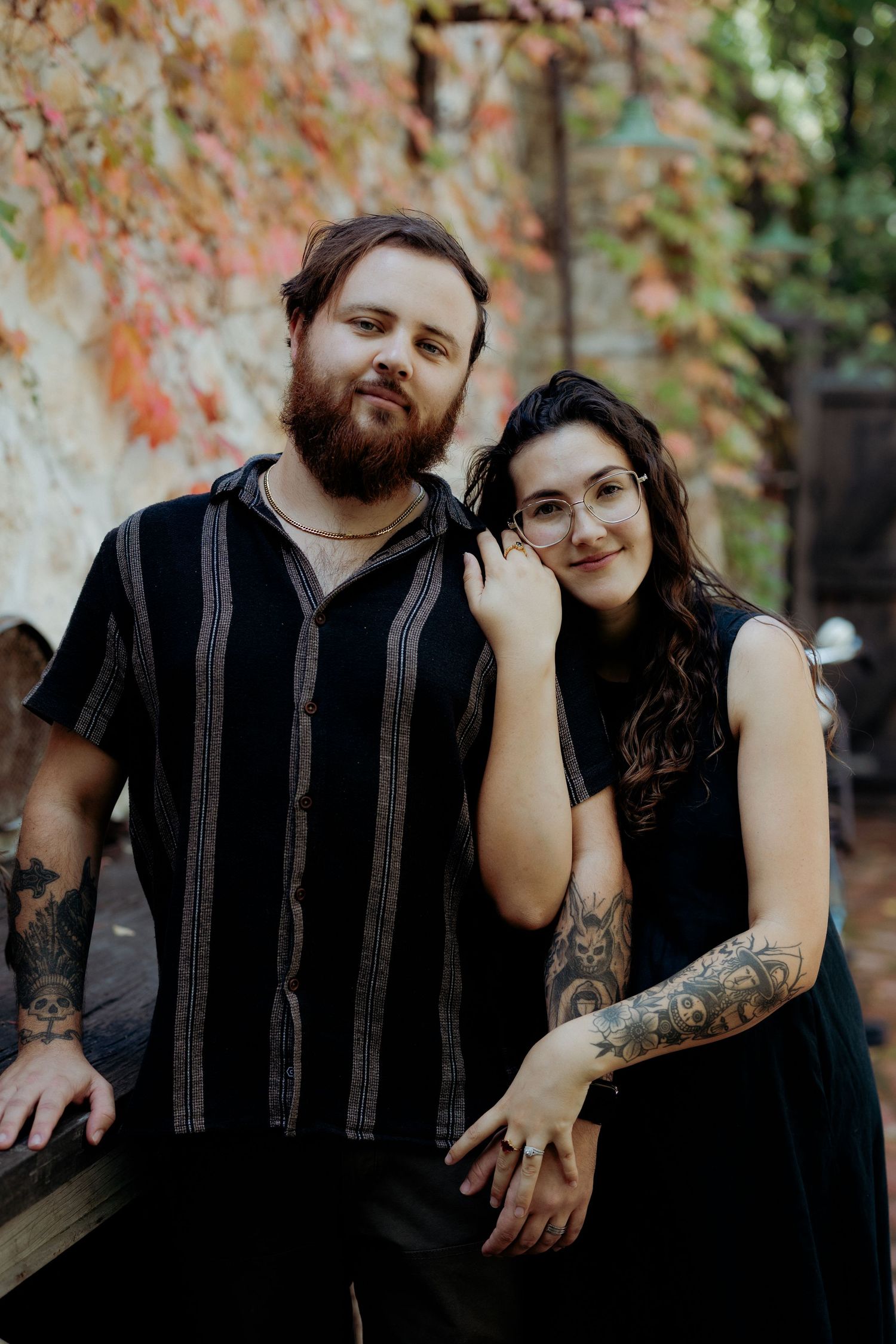 A tattooed couple in black outfits pose together outdoors with colorful autumn foliage in the background.