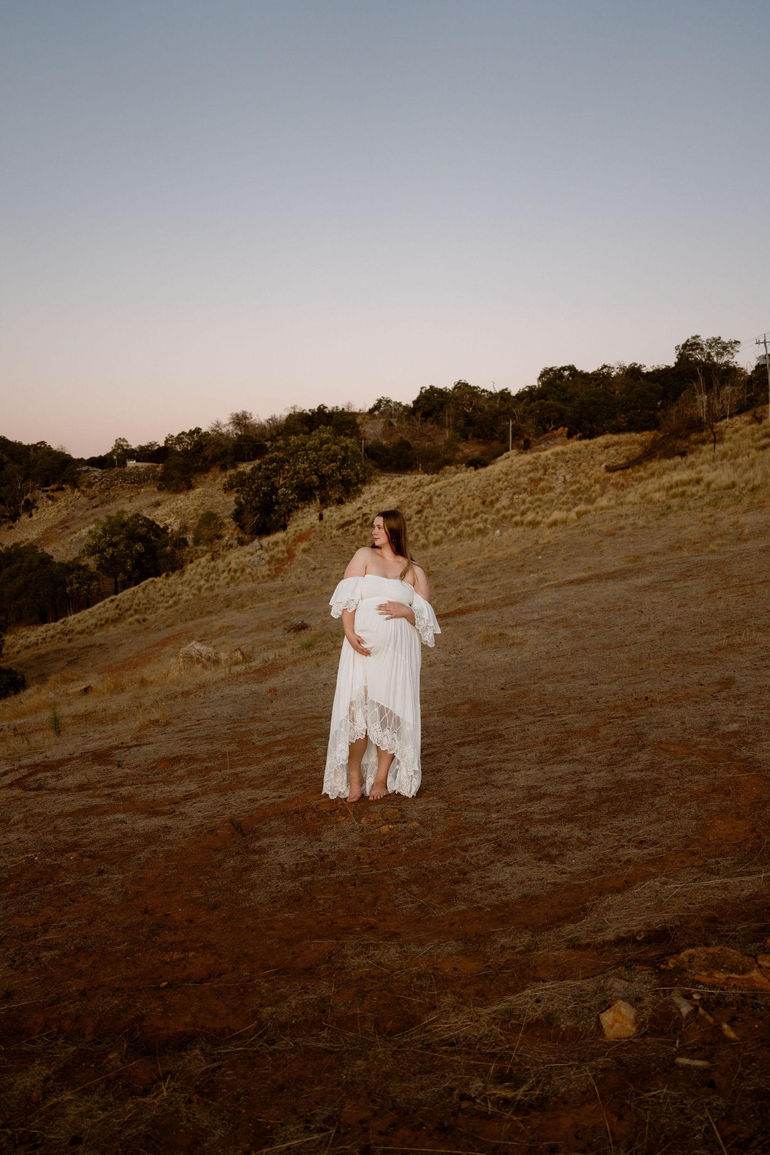A maternity photo of a woman in white dress standing on a hillside at sunset.