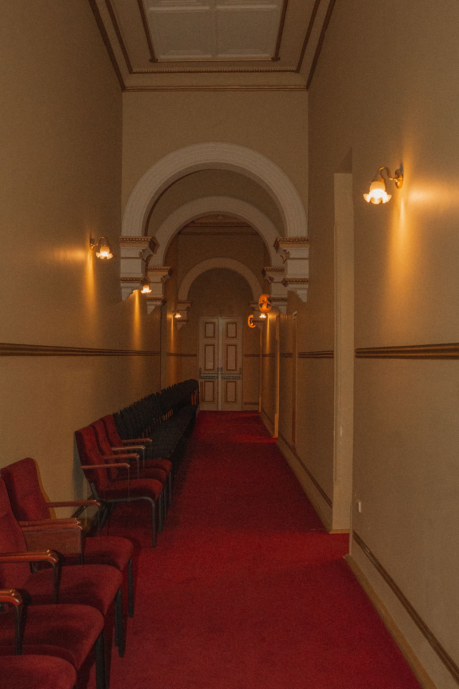 Elegant theater hallway with red carpet, vintage chairs along wall, and arched doorways illuminated by warm wall sconces.