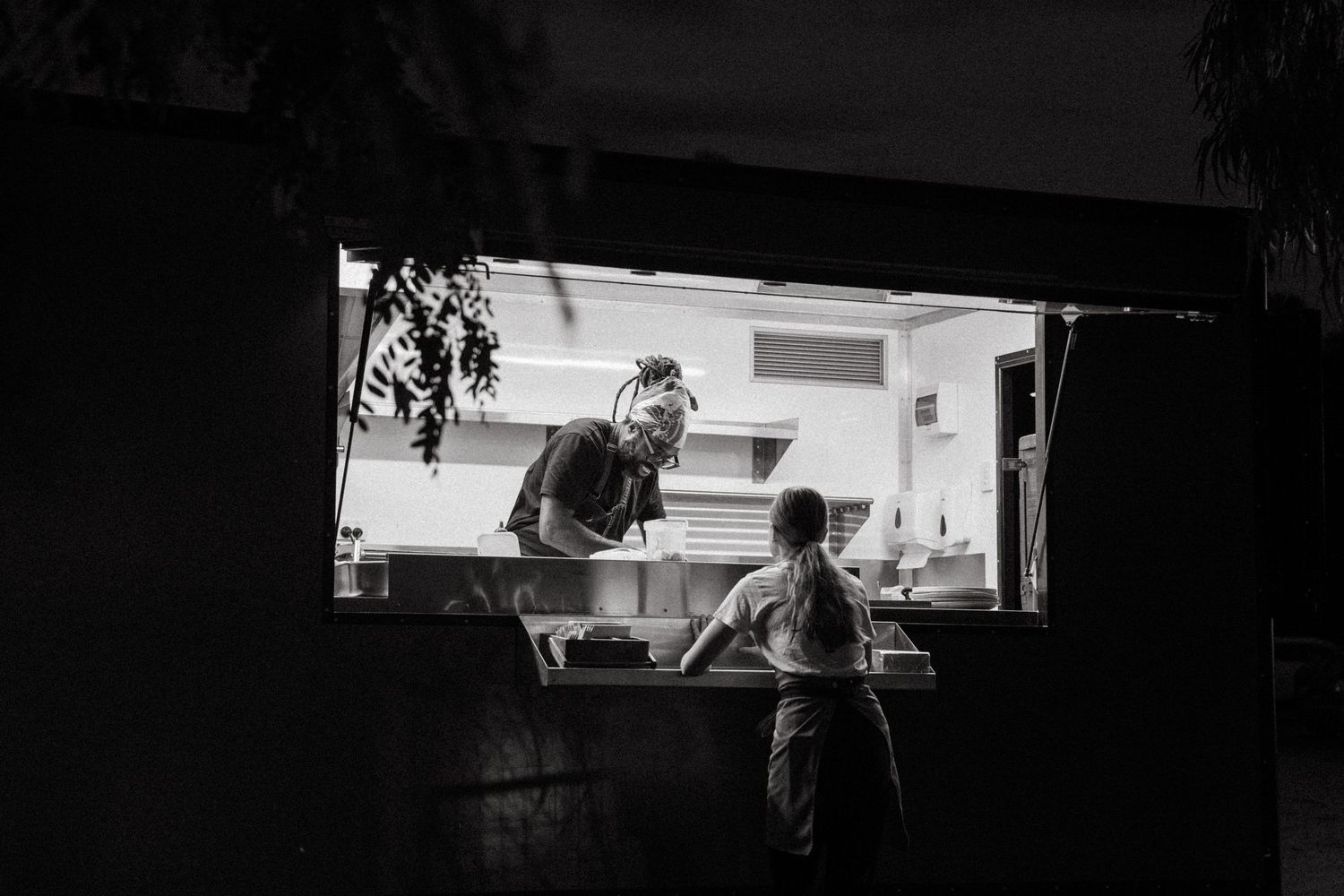 Black and white photo of a chef serving a customer through a food truck window at night.