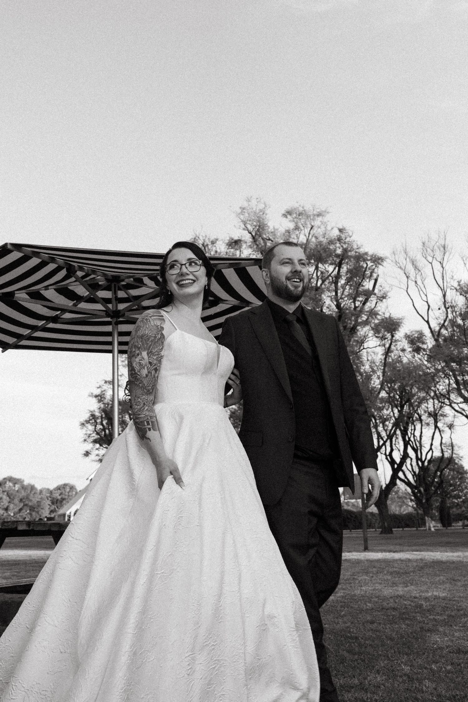 Black and white photo of a bride in a ball gown and tattooed groom in a suit walking outdoors near a striped umbrella.