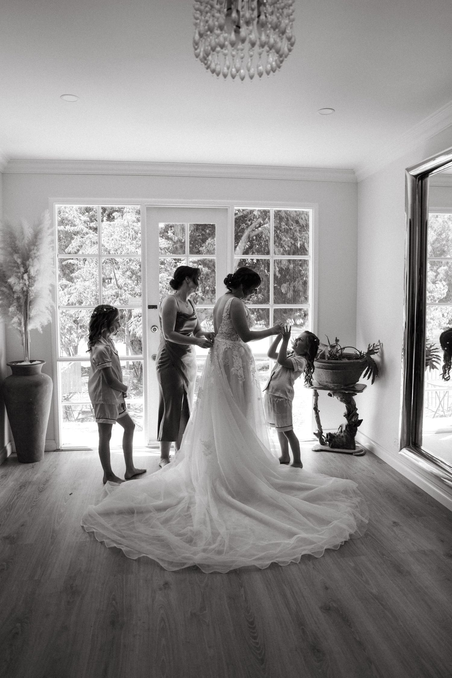 Black and white photo of a bride in a flowing gown with her bridesmaids and flower girls getting ready indoors.