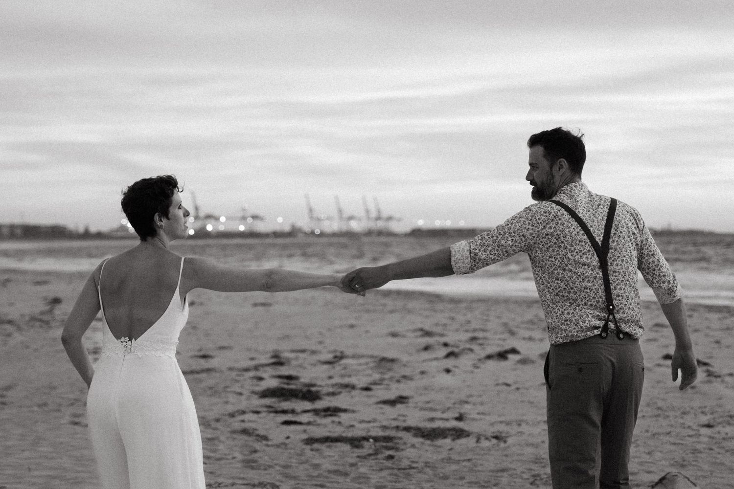 Black and white photo of a bride and groom holding hands on a beach, with industrial cranes visible in the background.