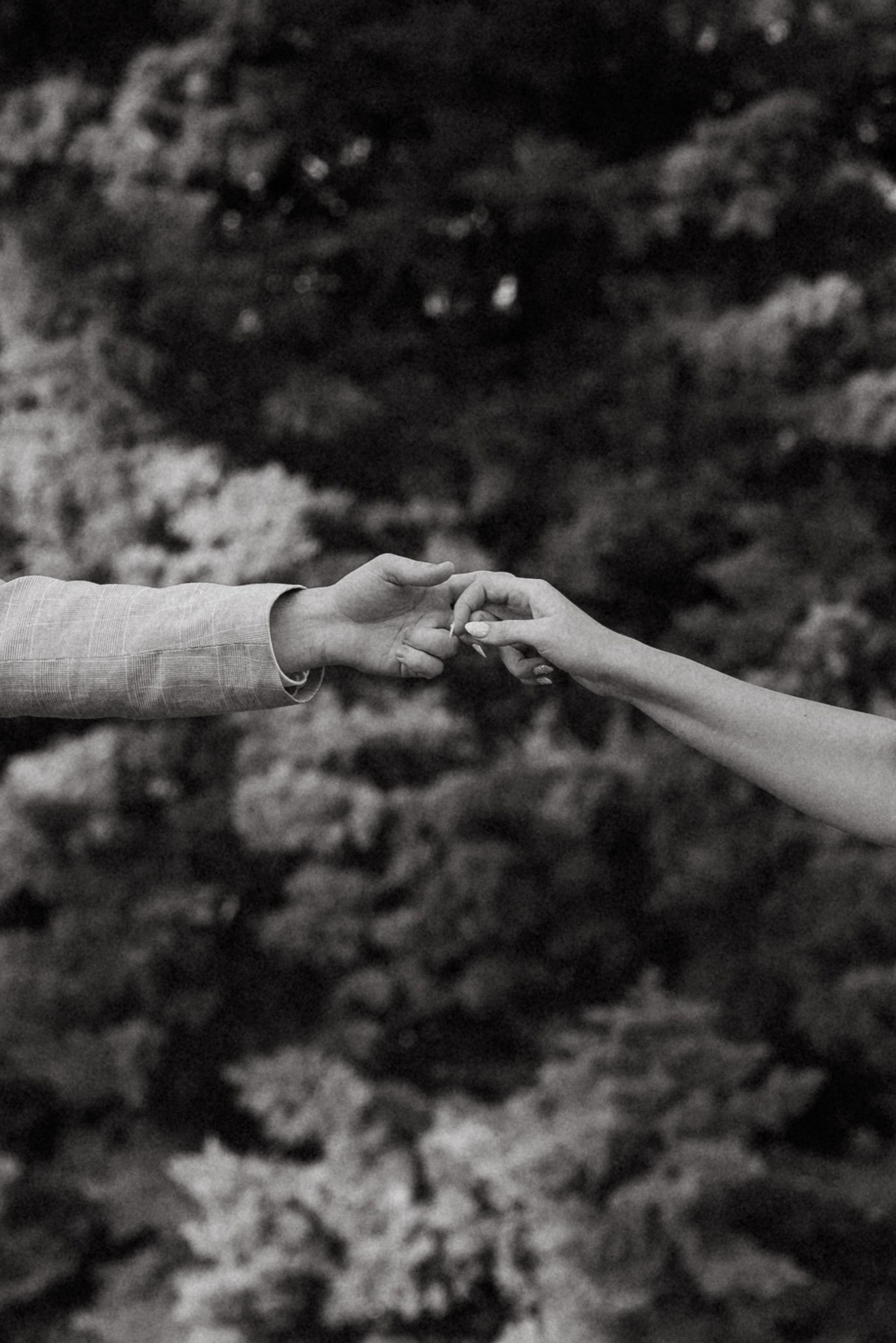 Black and white photo of two people gently touching fingertips with lush trees in the background.