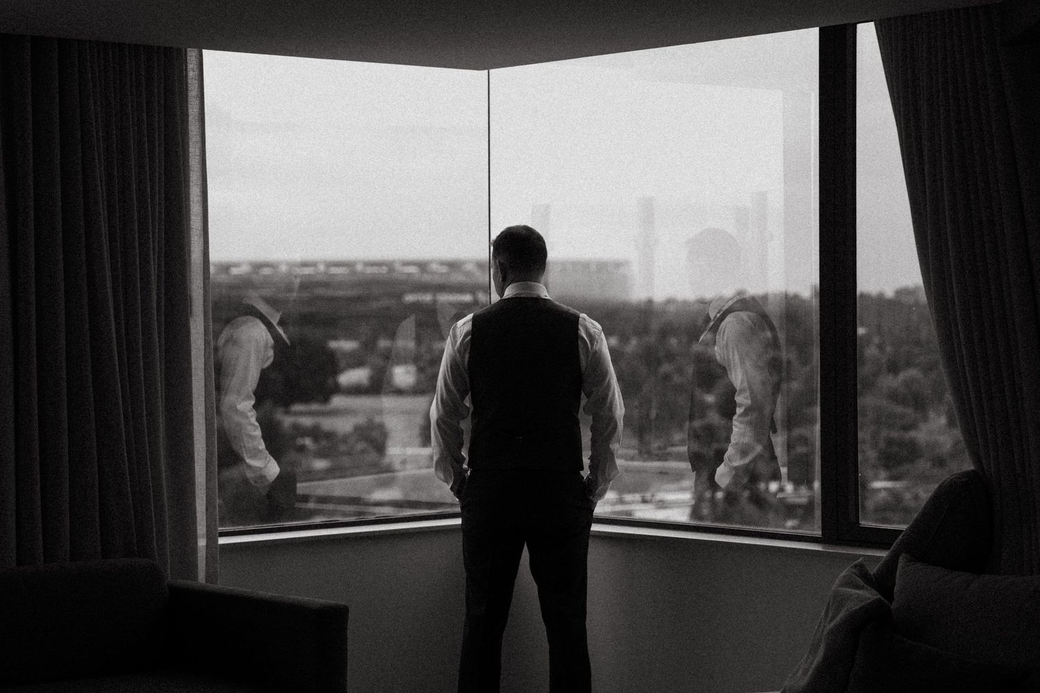 Man in vest stands before large corner windows overlooking city skyline in dramatic black and white photo.