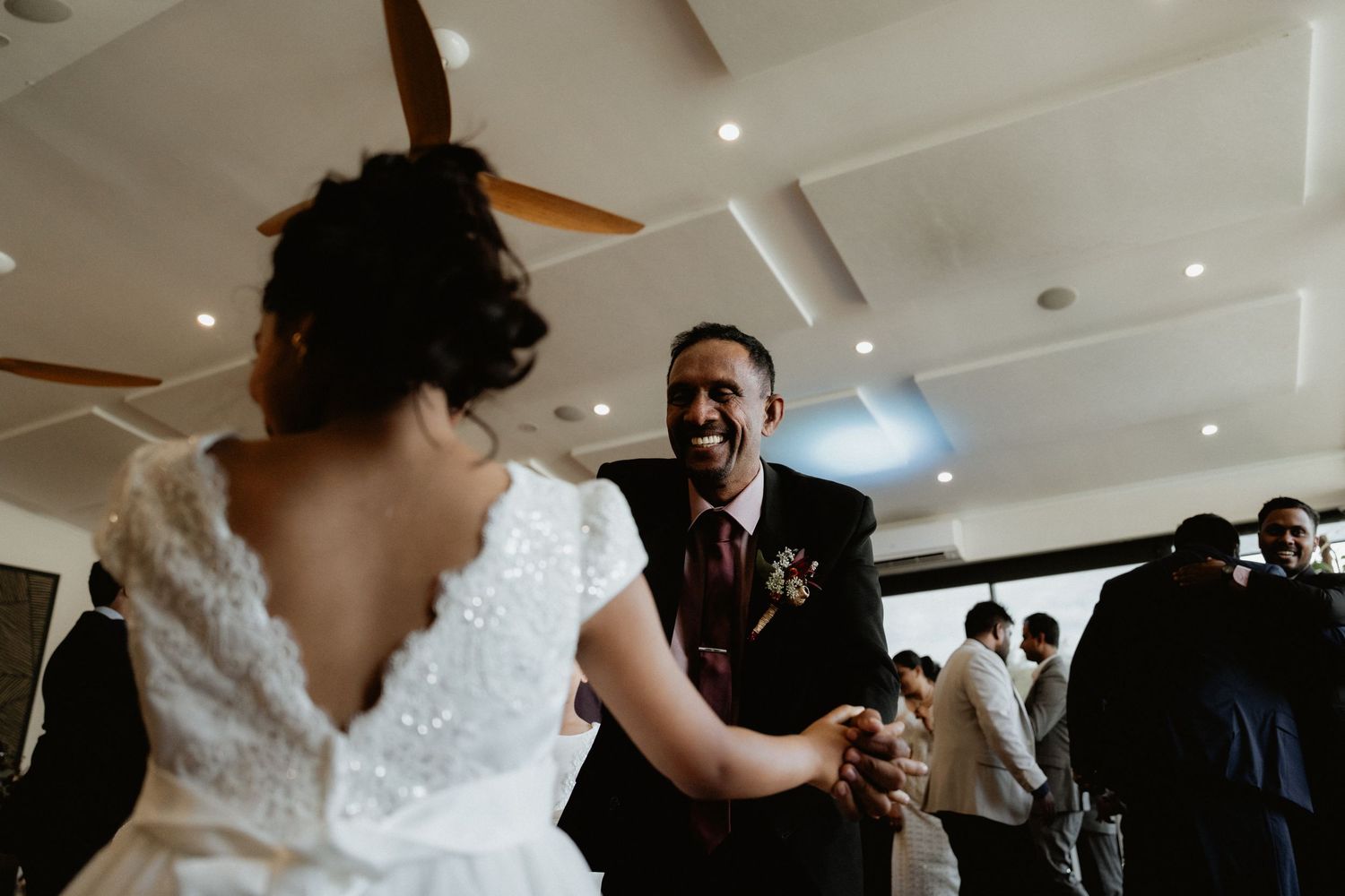 Joyful groom laughing while holding hands with bride in white lace wedding dress during reception dance.
