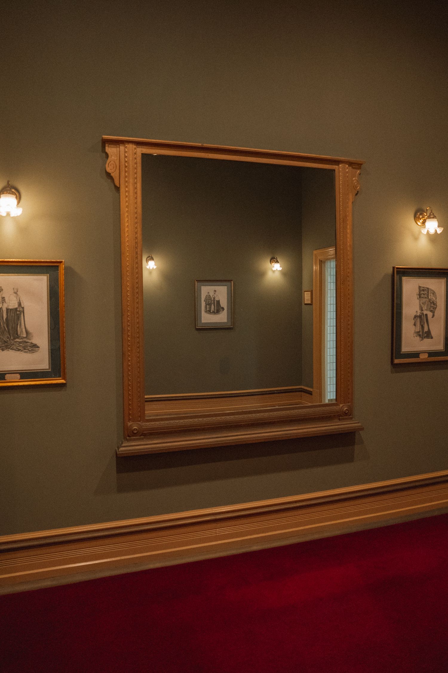 Elegant gallery hallway with warm lighting showcases framed artwork on sage green walls and rich red carpeting.