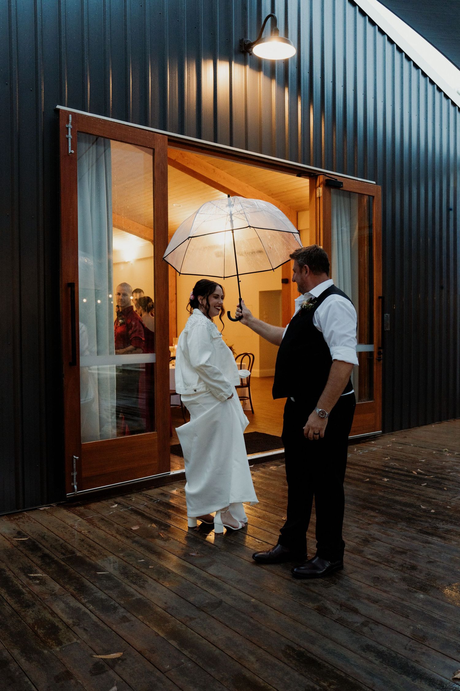 Bride and groom share a romantic rainy evening moment under a clear umbrella outside a warmly lit barn venue.