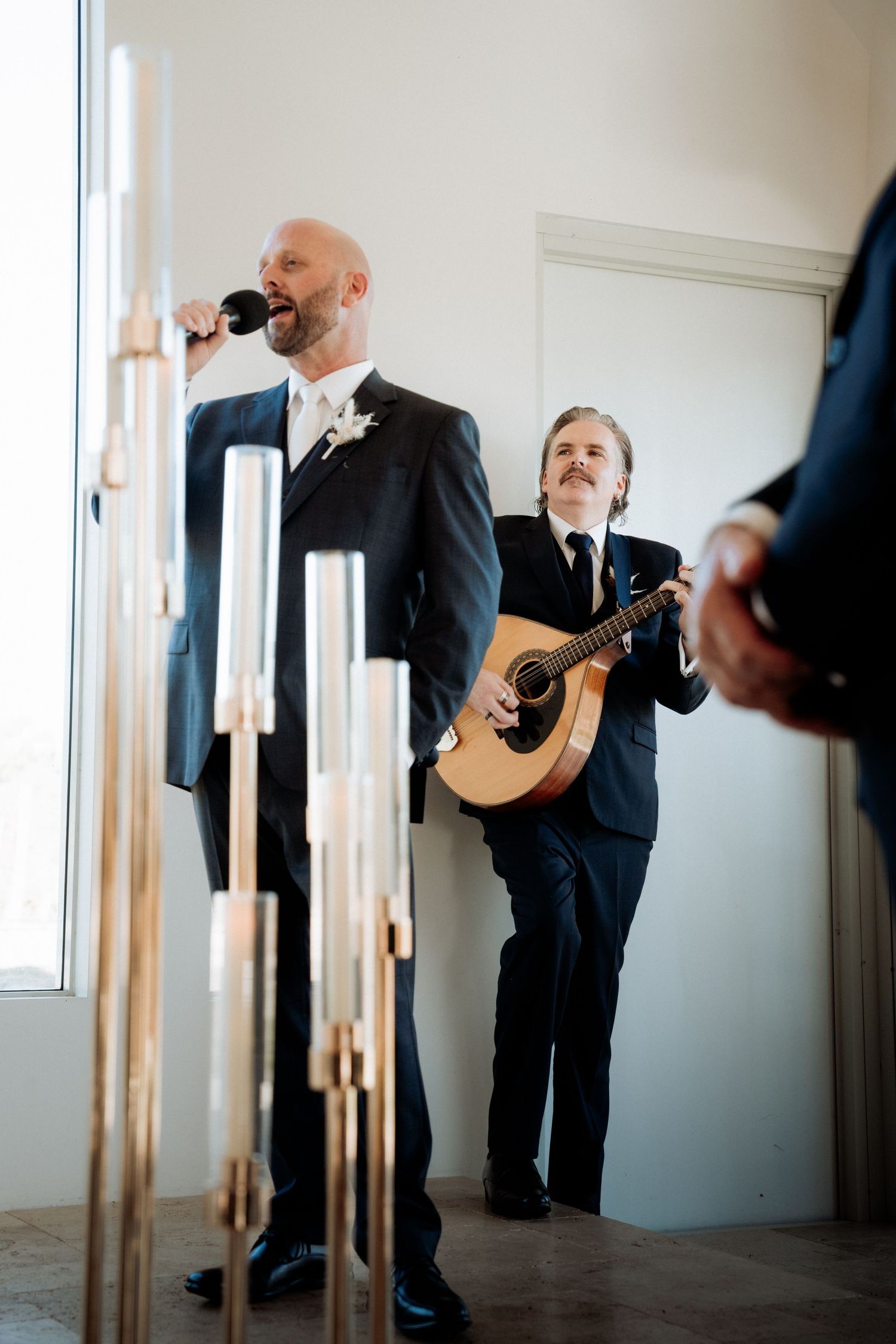 Two men in suits perform at a wedding, one singing into a microphone and one playing acoustic guitar near elegant candles.
