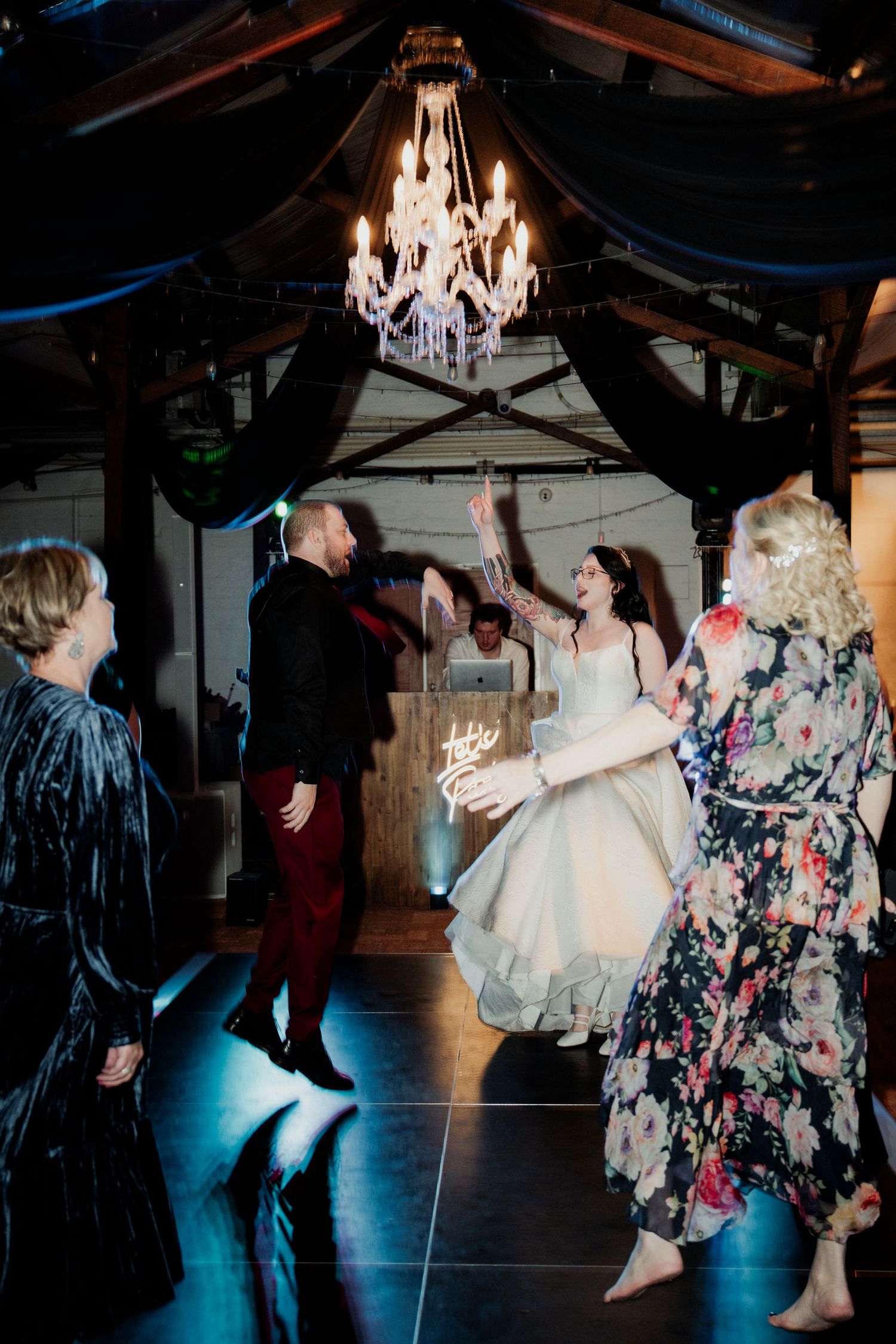 Bride in white gown dances joyfully with groom at wedding reception under elegant chandelier with guests.