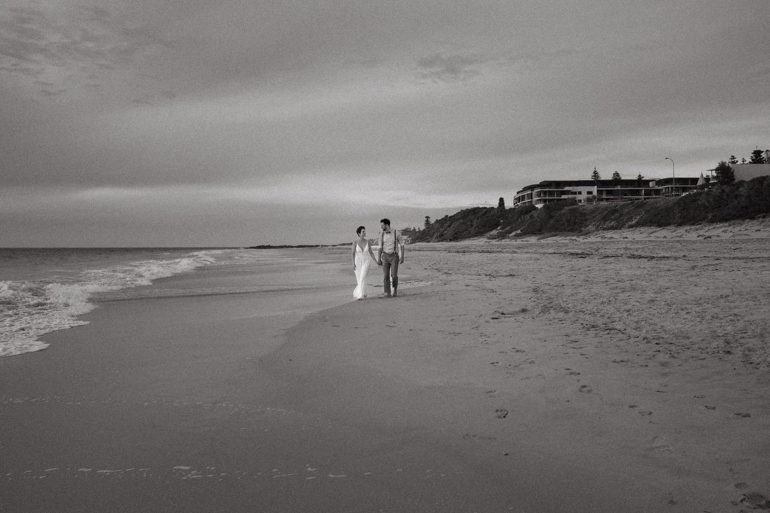 Black and white photo of a couple in wedding attire walking along a sandy beach with ocean waves and cloudy sky.