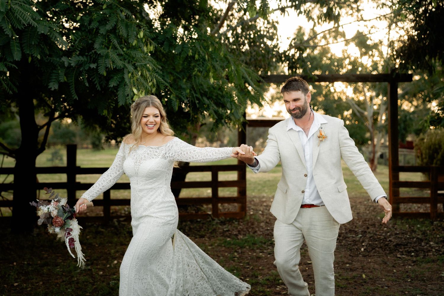 Bride and groom holding hands and smiling outdoors near a wooden fence and lush green trees on their wedding day.