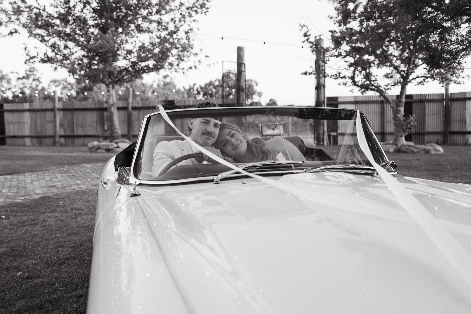 Black and white photo of a bride and groom smiling in a classic white convertible car on their wedding day.