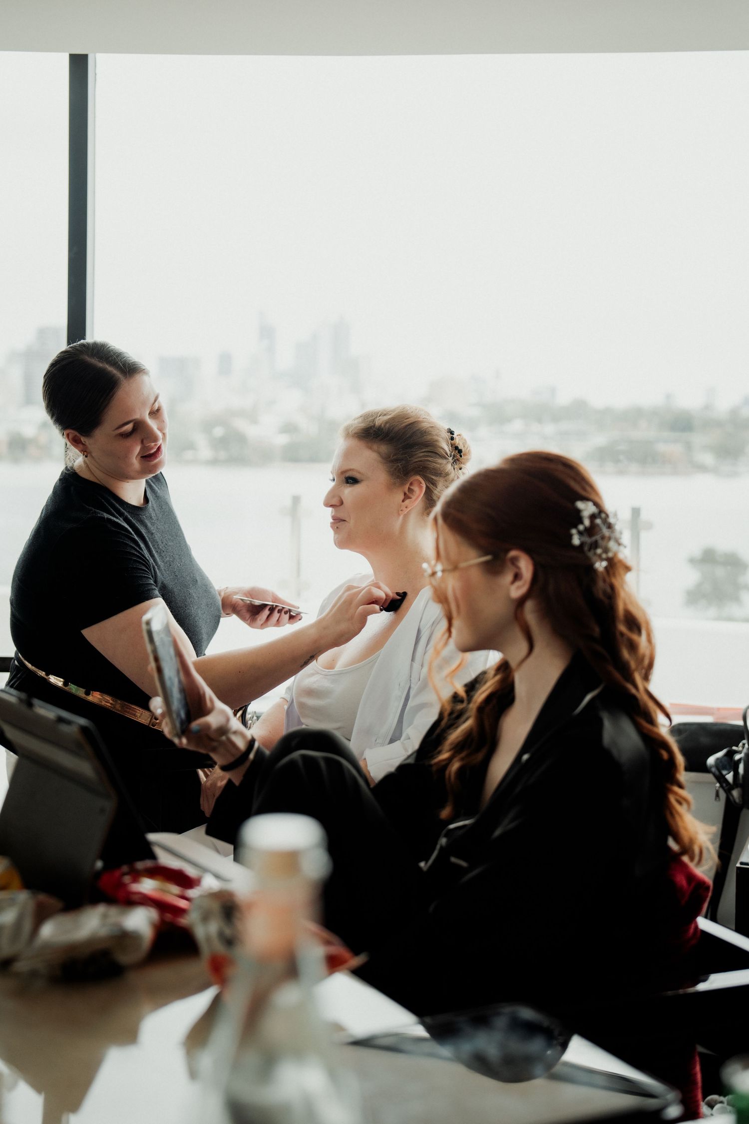 Makeup artist applying makeup to bride in white robe, with bridesmaid in black satin robe, city skyline view.
