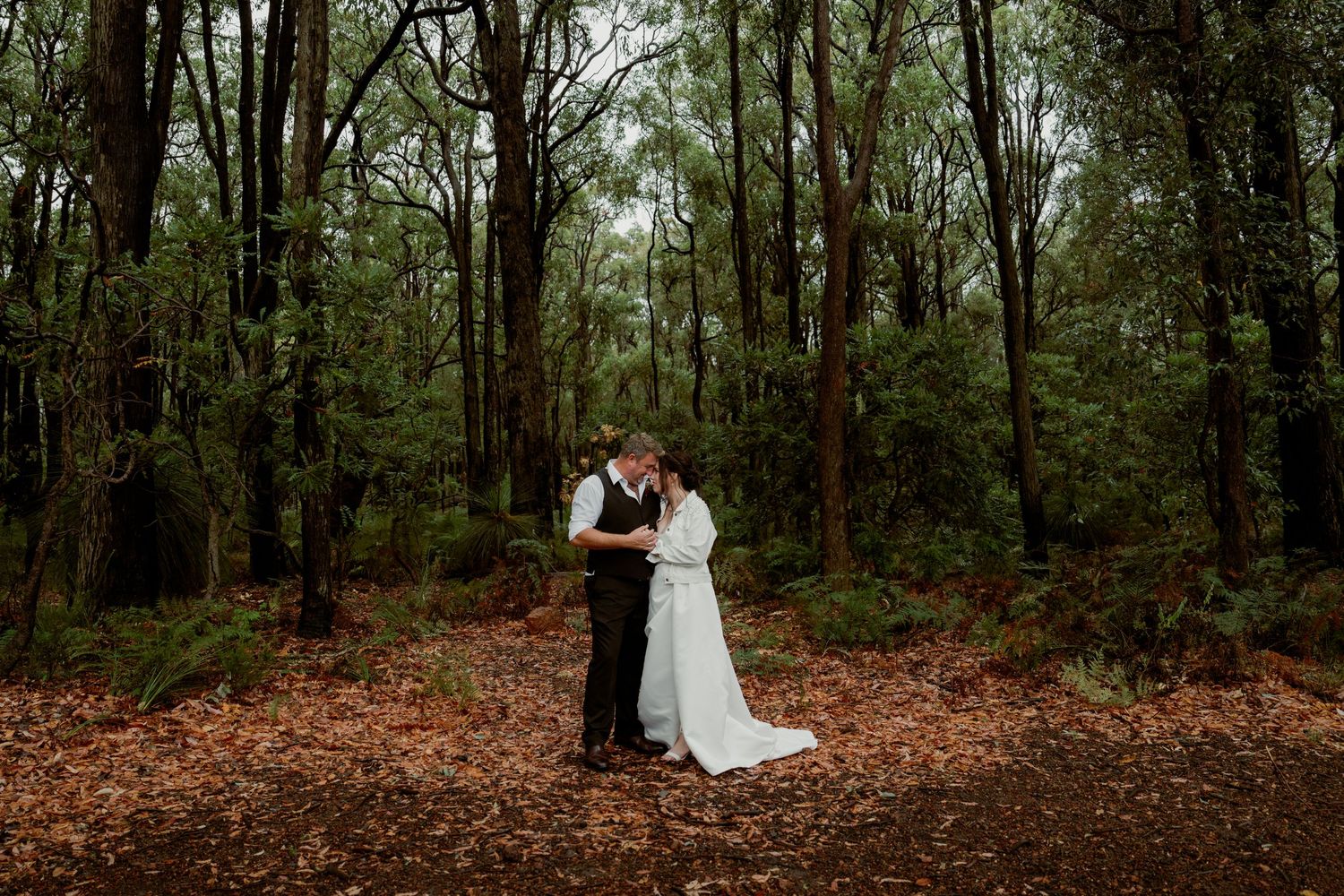 Bride and groom share a romantic kiss surrounded by tall trees in a lush forest setting on their wedding day.