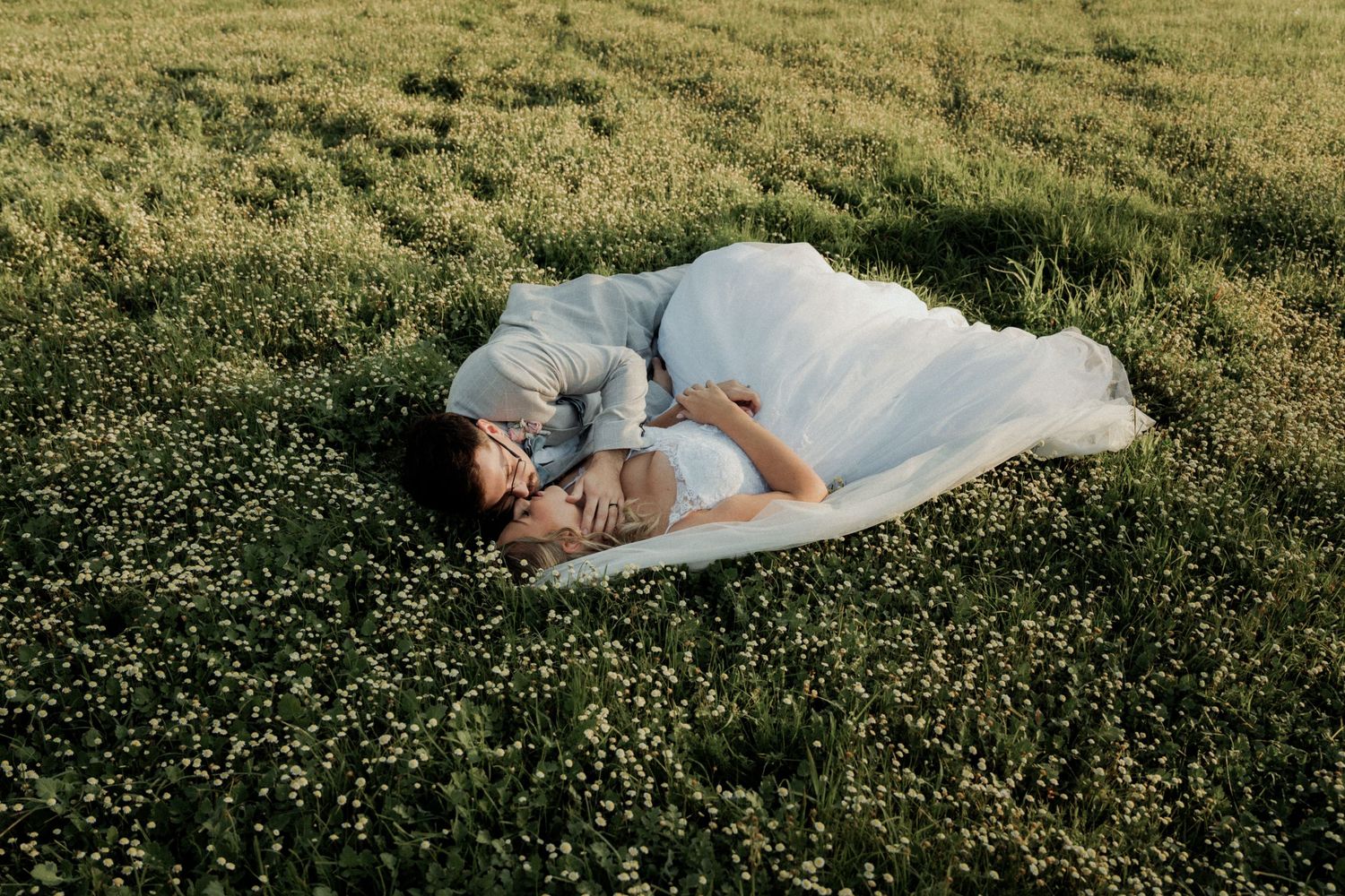 A bride in a white wedding gown lies peacefully in a lush green meadow filled with small wildflowers.