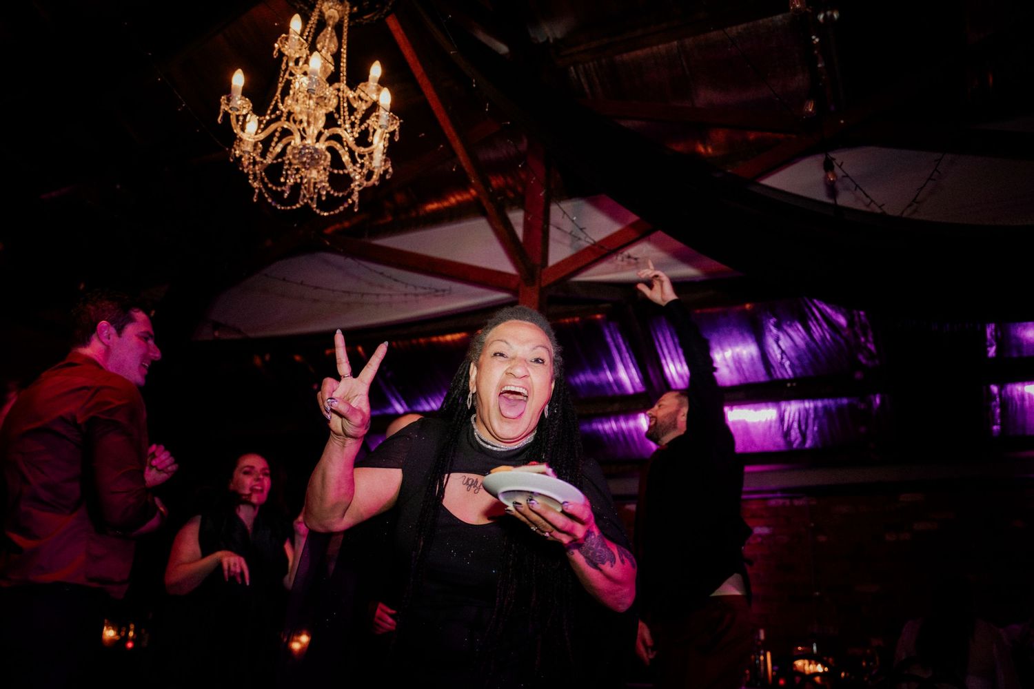 Woman flashing peace sign at lively indoor party with chandelier and purple lighting in background.
